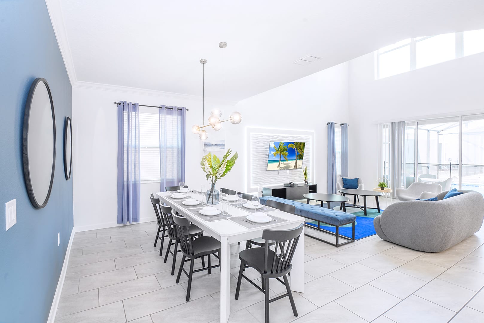 Open-concept living and dining area featuring a long white dining table with black chairs under a modern chandelier, and a living space with a gray sofa and blue accents, all set against white walls and a blue accent wall.