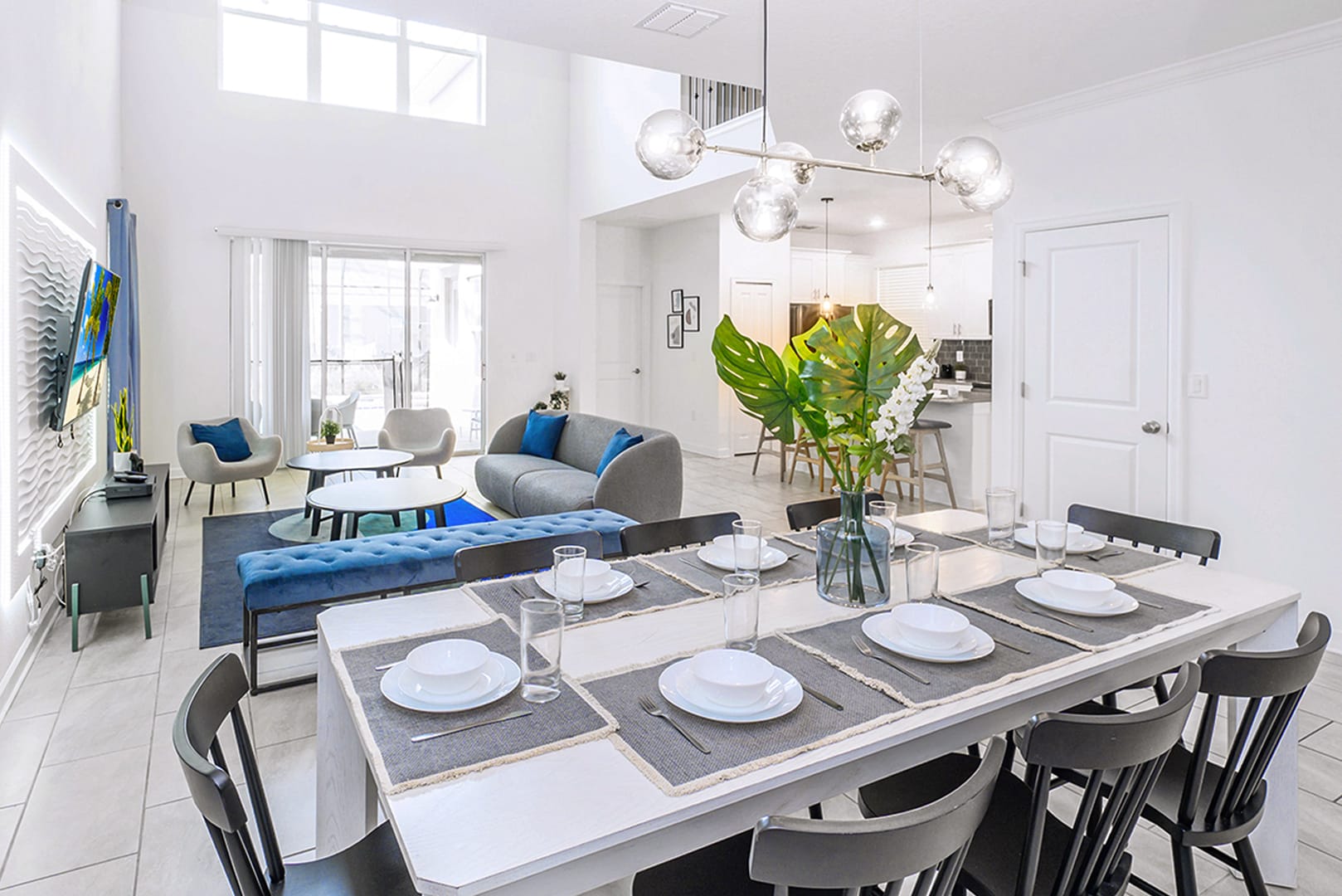 Expansive open-concept space with a white dining table set for eight, adorned with a large plant centerpiece, flowing into a bright living area with a gray sofa and blue accents, all under high ceilings with large windows.