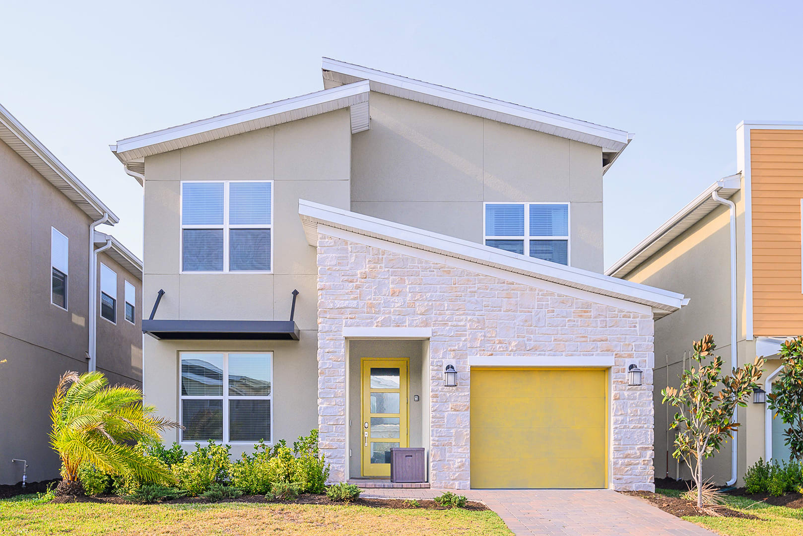 The modern two-story house features a light beige stucco exterior with white stone accents, a bright yellow front door, and a matching yellow garage door, surrounded by green landscaping.