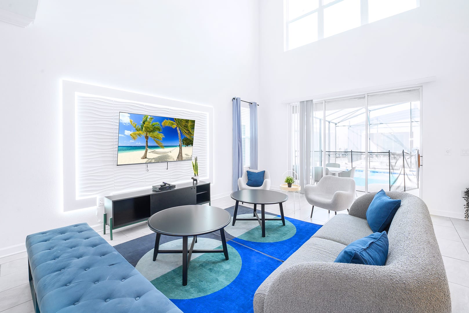 Bright, modern living room with white walls, high ceilings, and a large gray curved sofa with blue pillows, facing a TV mounted on a textured white wall with LED backlighting. A blue and teal geometric rug anchors two black round coffee tables.