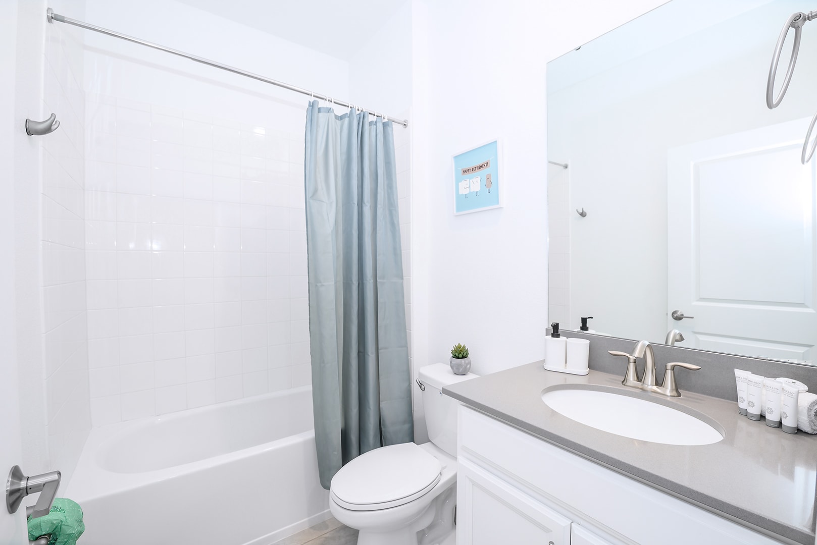 Bright white bathroom with a bathtub/shower combo, a light blue shower curtain, and a single vanity with a gray countertop and a large mirror. A small plant and toiletries are on the counter.