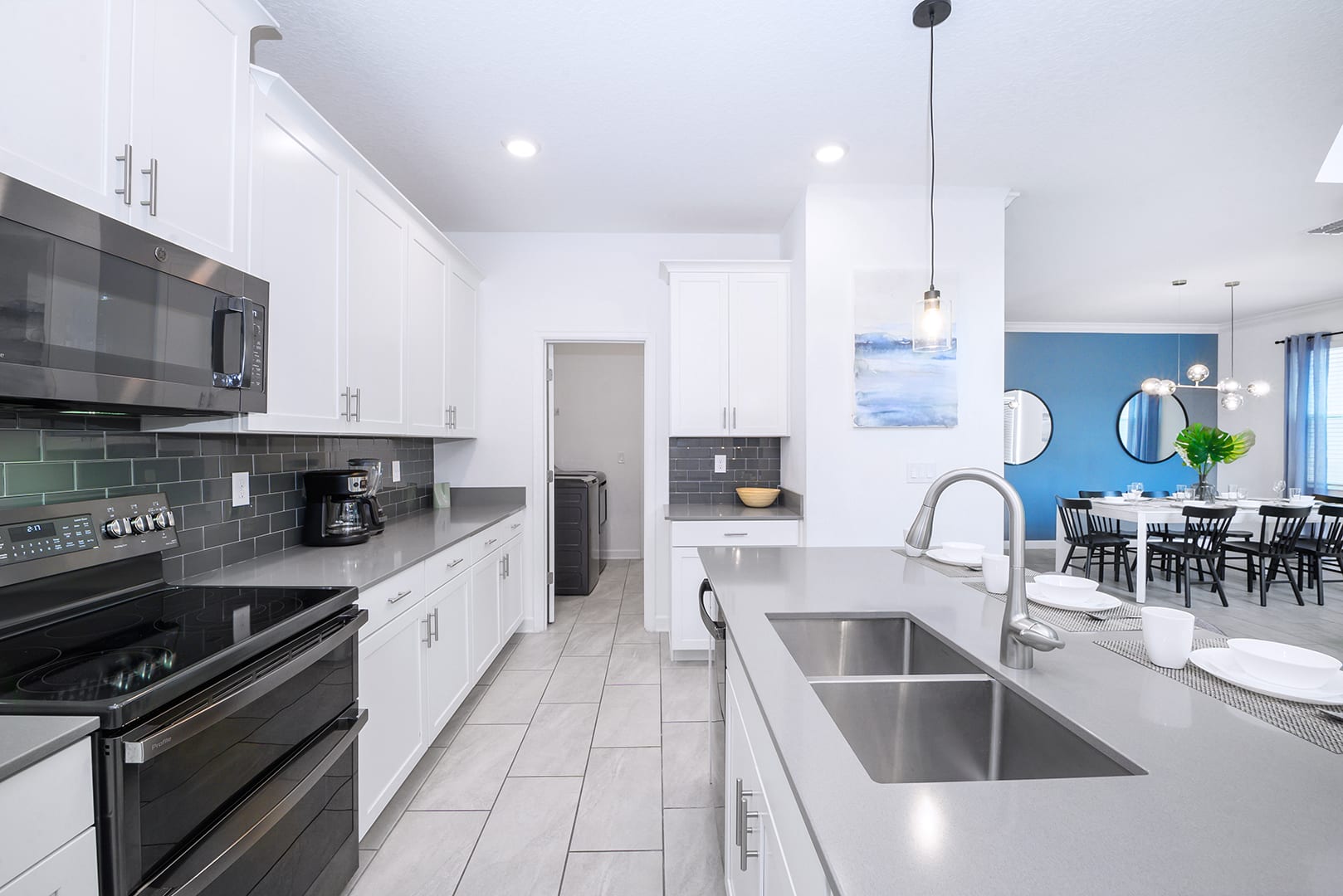 Galley-style kitchen with white upper and lower cabinets, dark gray subway tile backsplash, and light gray countertops, featuring a black stove, microwave, and coffee maker, with a laundry area visible beyond.