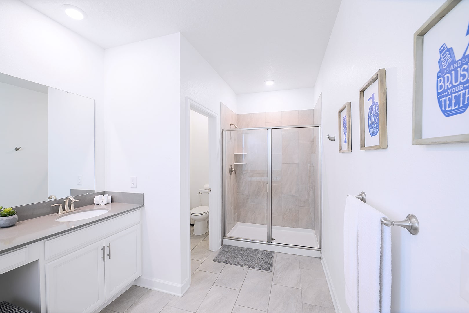 Clean, bright bathroom with a walk-in shower featuring glass doors and tiled walls, a toilet in a separate alcove, and a single vanity with a sink and mirror, decorated with framed wall art.