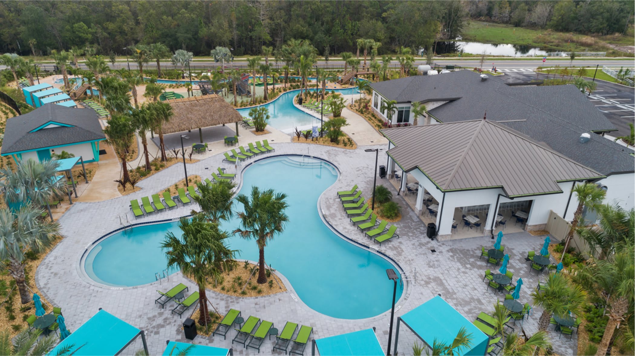 Aerial view of a large communal pool complex with multiple interconnected pools, a lazy river, numerous lounge chairs, and several buildings including a thatched-roof cabana, surrounded by lush landscaping.