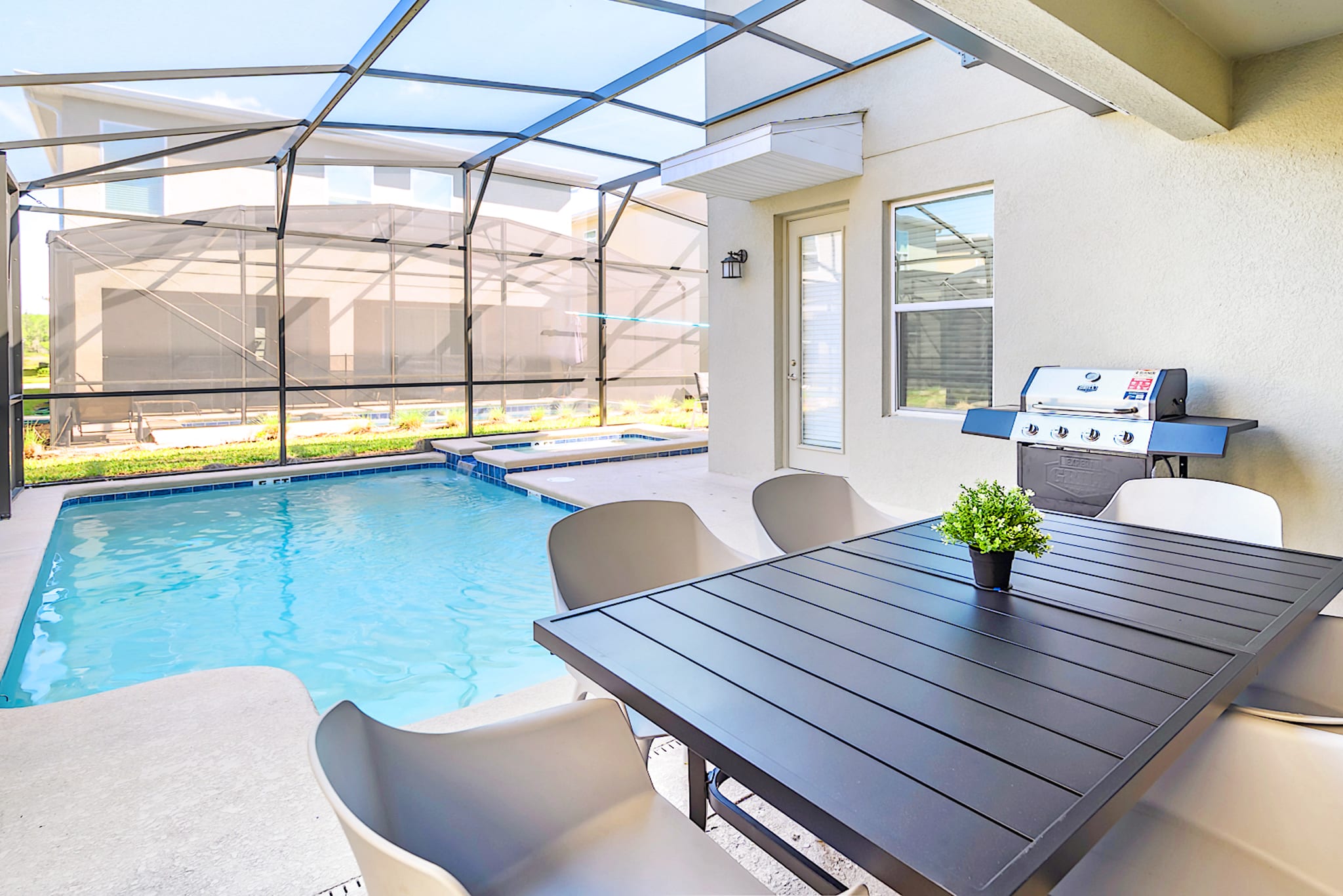 A screened-in outdoor patio features a rectangular swimming pool with an integrated hot tub, a dark dining table with four chairs, and a gas grill, all under a clear roof.