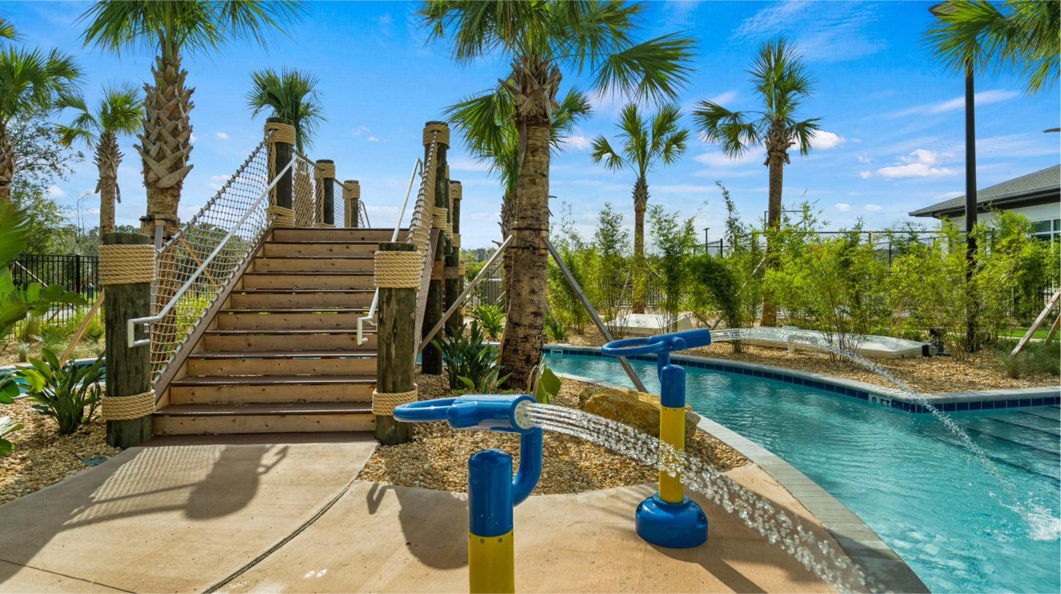 Water play area featuring a shallow lazy river with water sprayers and a wooden bridge structure with rope railings, surrounded by lush tropical landscaping and palm trees under a bright blue sky.