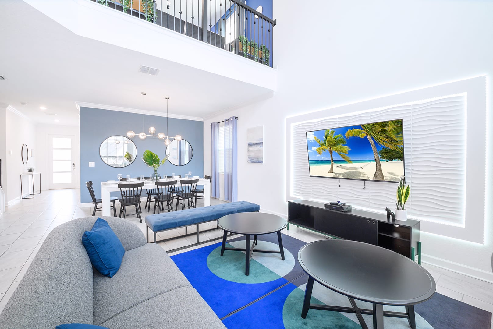 The modern living room features a gray sofa, a blue and teal geometric rug, and a large flat-screen TV mounted on a textured white accent wall with LED backlighting.