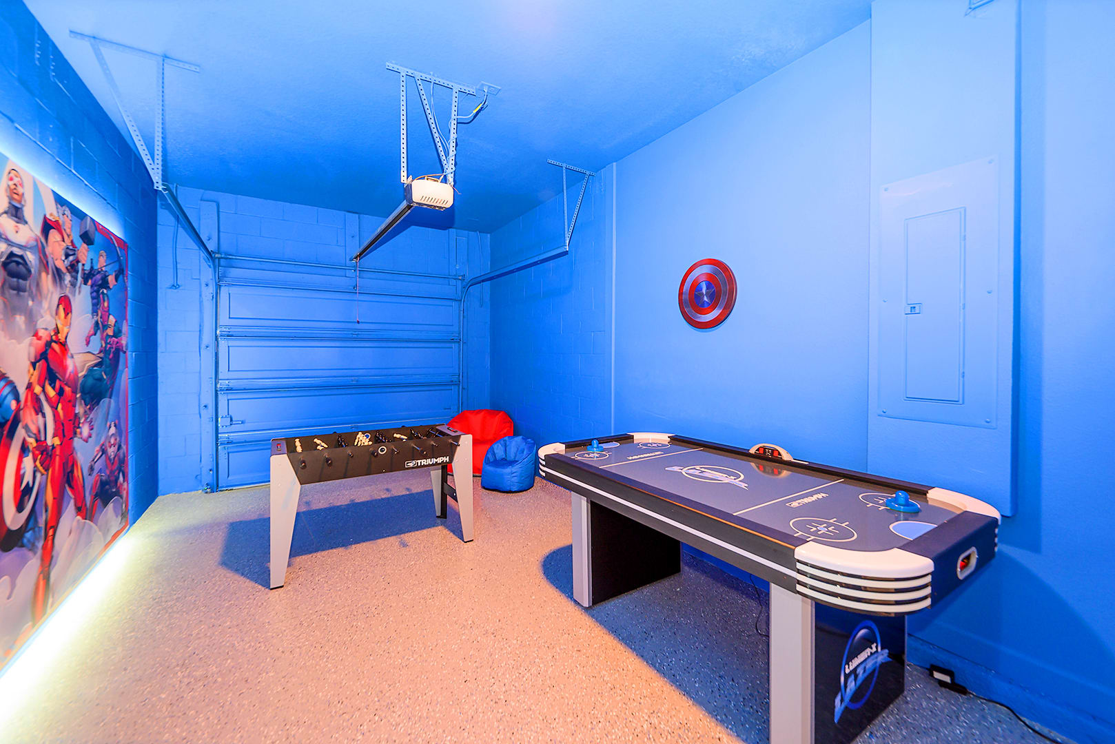 A vibrant blue-painted garage converted into a game room features a foosball table, an air hockey table, and a large backlit Marvel superhero mural, with a Captain America shield on the wall.