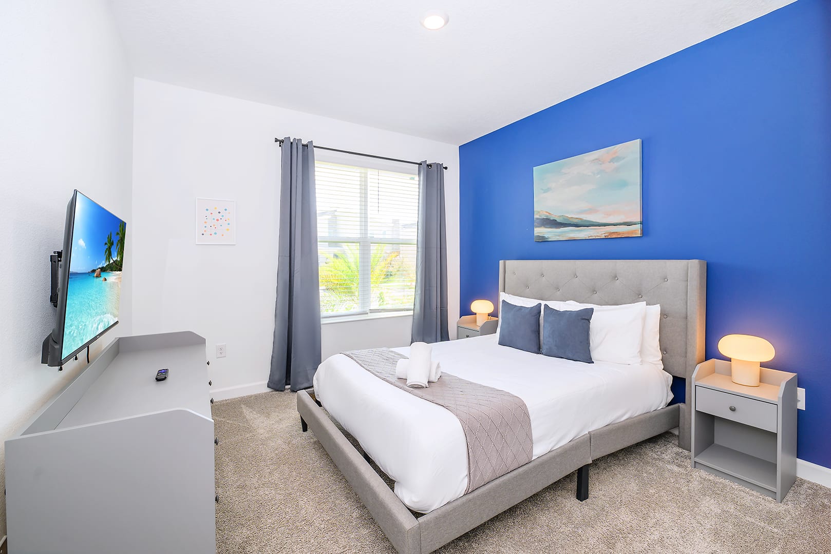 Modern bedroom featuring a gray upholstered bed with white bedding, a blue accent wall with abstract art, and a wall-mounted TV opposite the bed. Two bedside tables with lamps flank the bed.