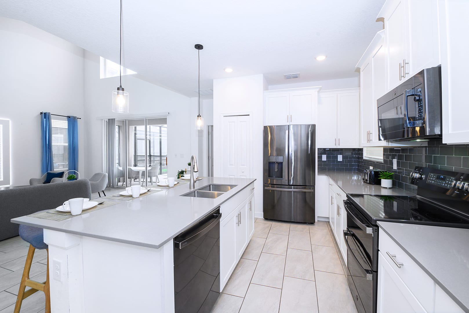 Bright, open-plan kitchen with white cabinetry, light gray countertops, and stainless steel appliances, including a large island with a double sink and dishwasher, connecting to the living area with views of the outdoor pool.