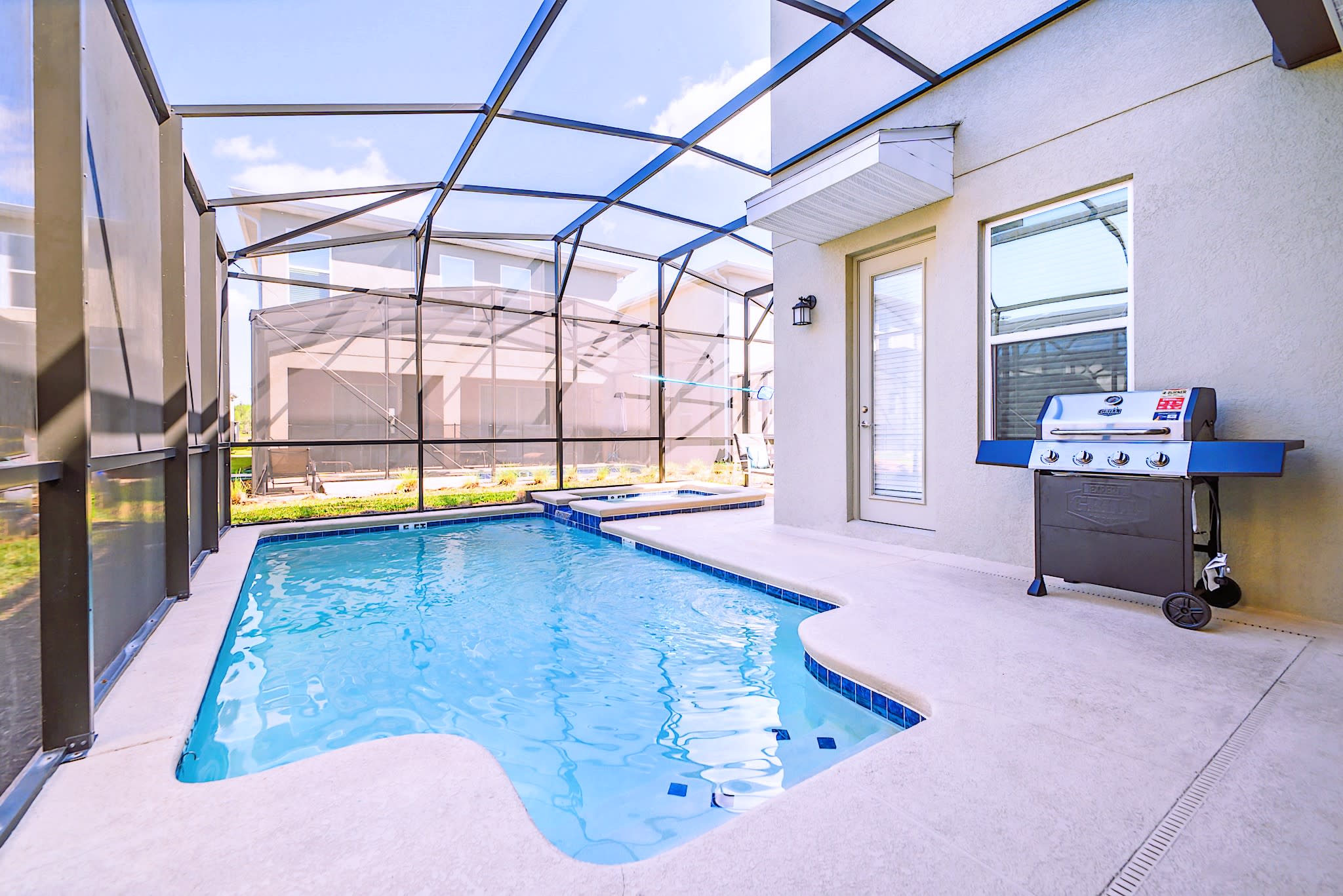 A private screened-in pool area features a light blue swimming pool with an integrated hot tub, a gas grill, and a view of neighboring houses through the screen enclosure.