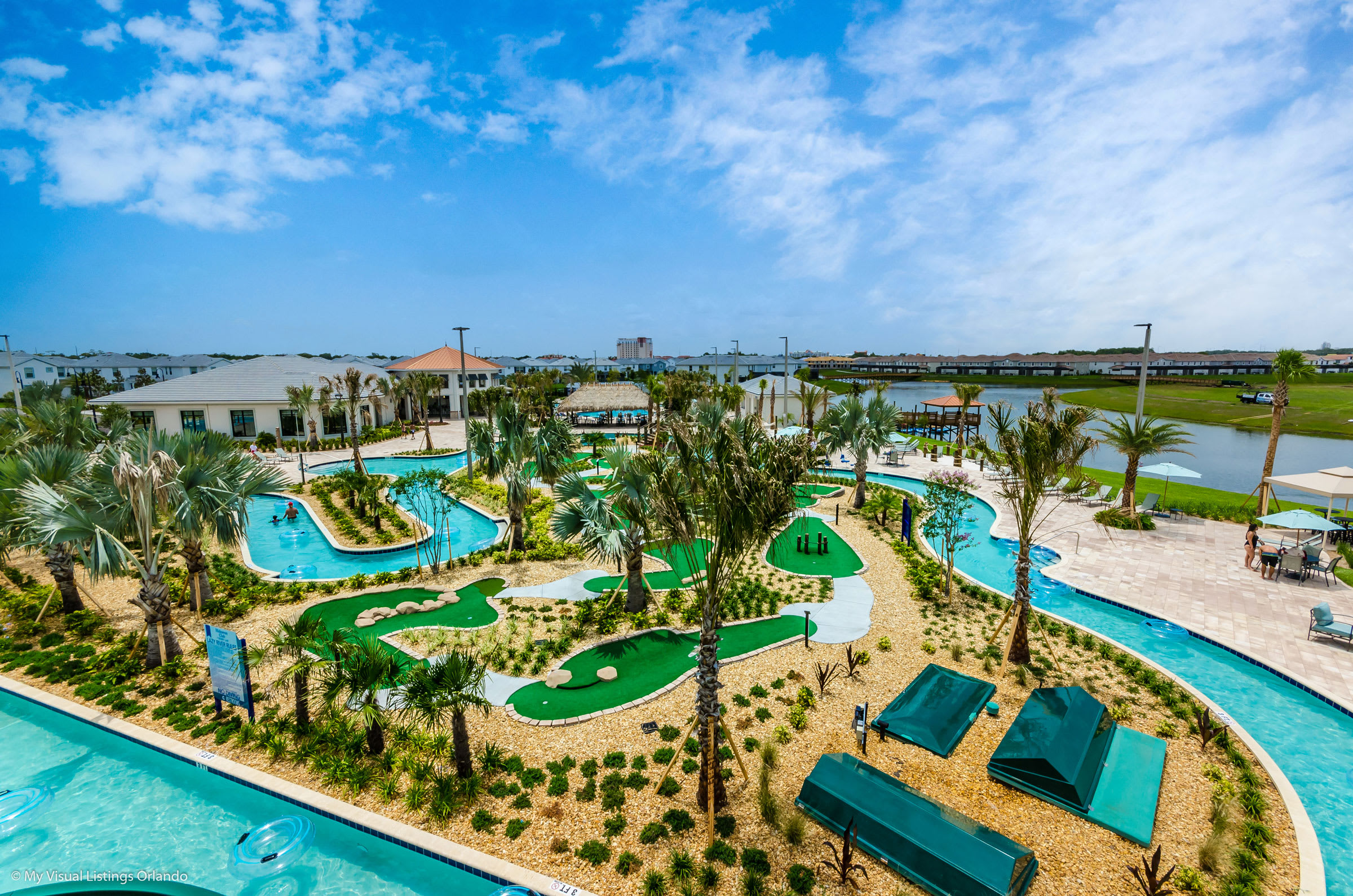 Expansive aerial view of a resort-style recreation area featuring a winding lazy river, miniature golf course, multiple pools, and various lounge areas, surrounded by palm trees and residential buildings.