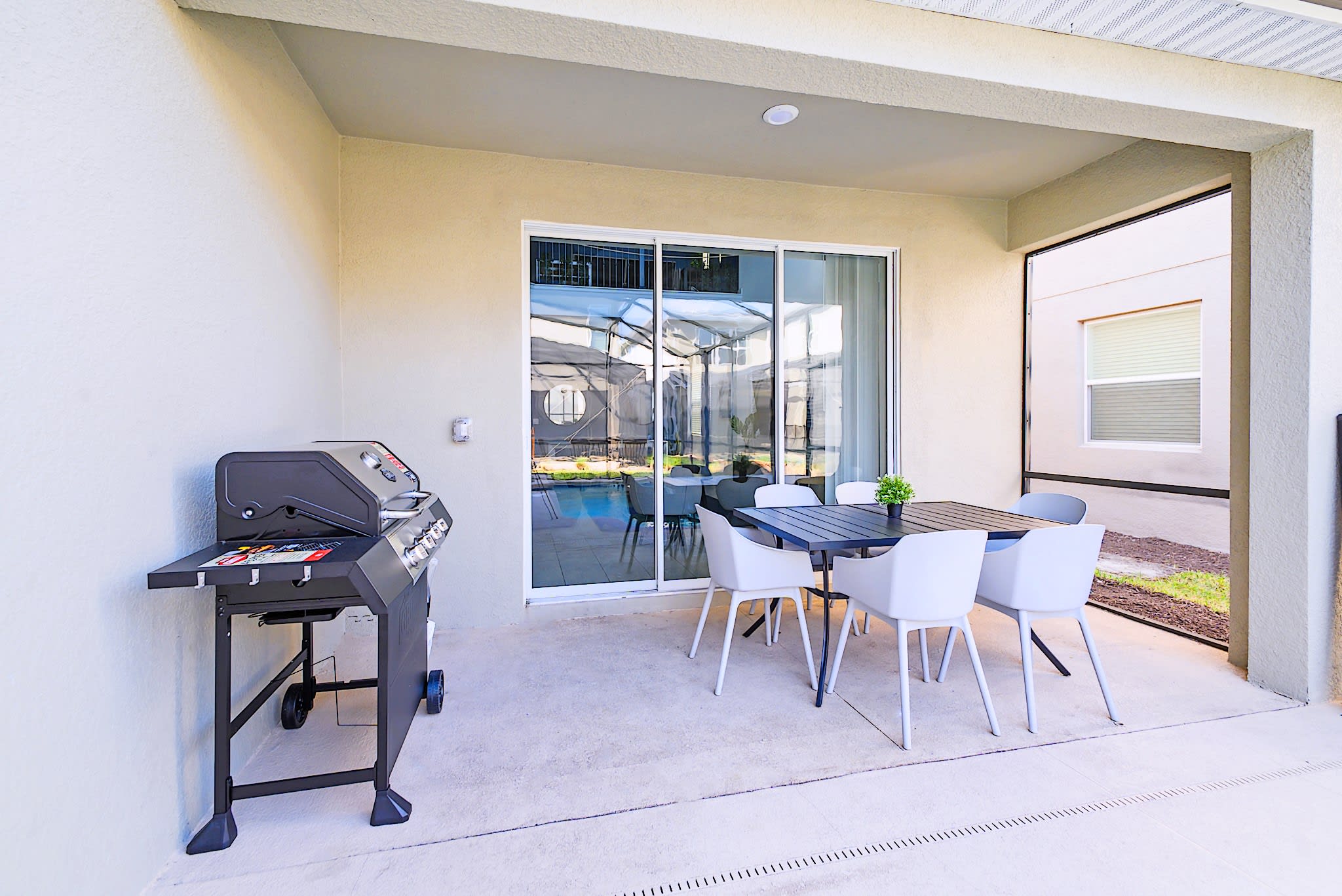 A covered outdoor patio features a dark dining table with four white chairs and a black gas grill, with a sliding glass door leading into the house and a view of the screened pool area.