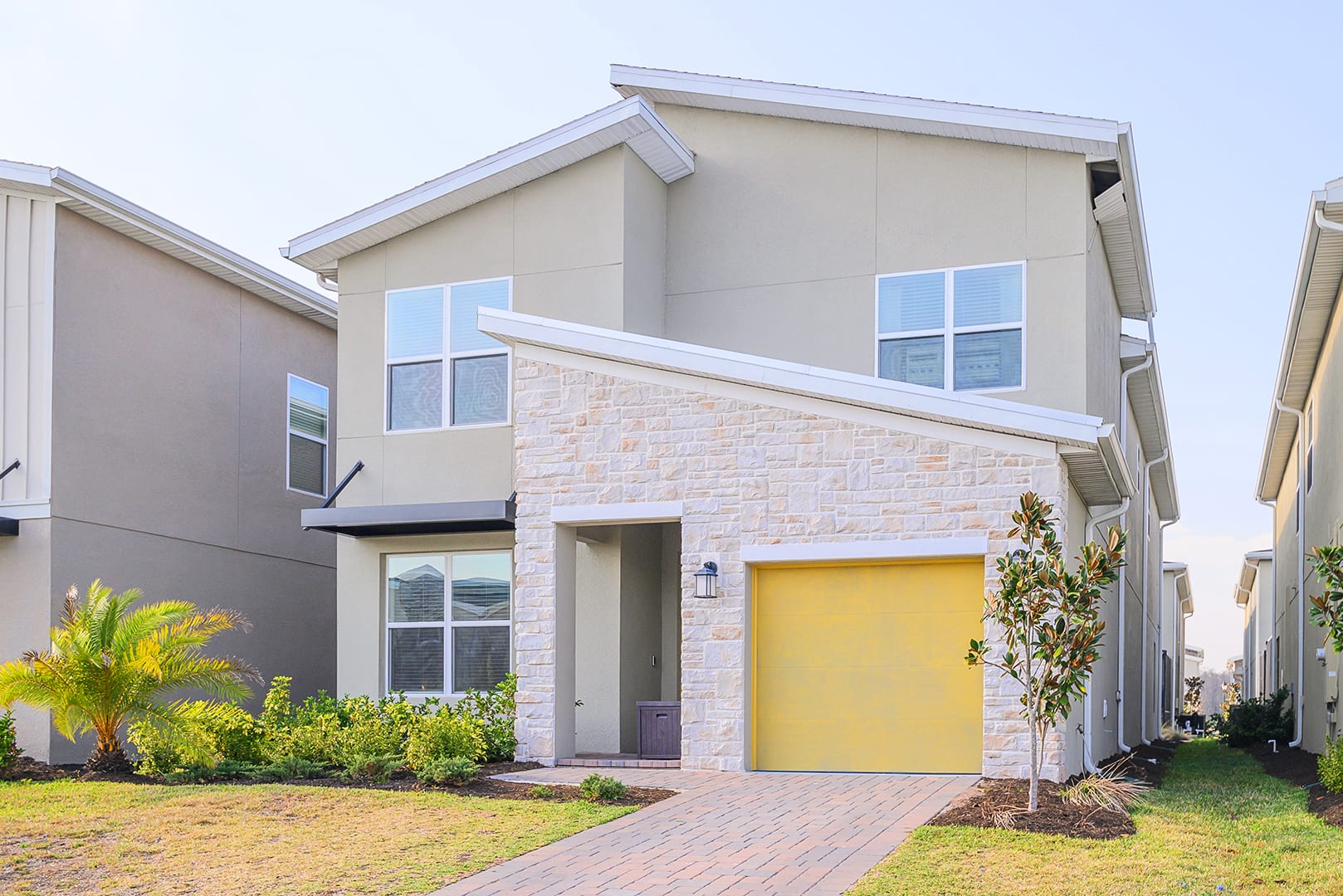 Modern two-story house with light beige stucco and a prominent stone facade on the lower level, featuring a bright yellow garage door and a paved driveway leading to the entrance.