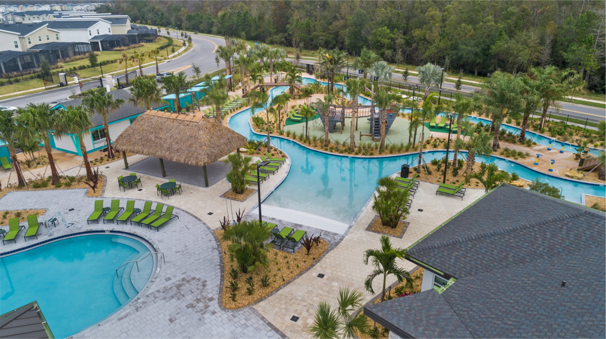 Aerial view of a vibrant resort-style pool area featuring a lazy river, multiple swimming pools, a water playground, and a thatched-roof cabana, all set amidst palm trees and green lounge chairs.