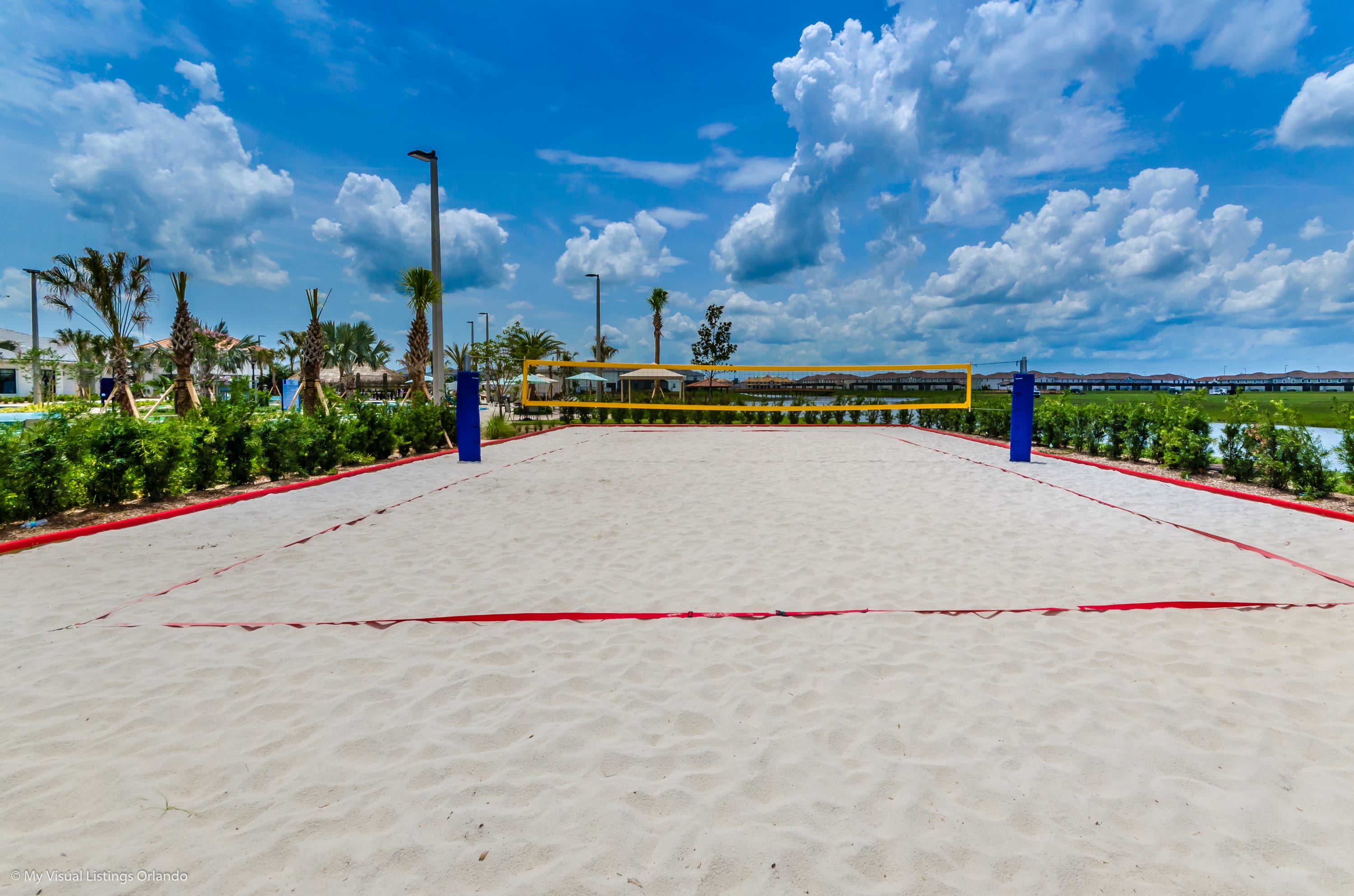A pristine outdoor sand volleyball court with a yellow net and red boundary lines is set against a backdrop of palm trees and a distant body of water. The bright blue sky with white clouds suggests a sunny day, ideal for outdoor activities.