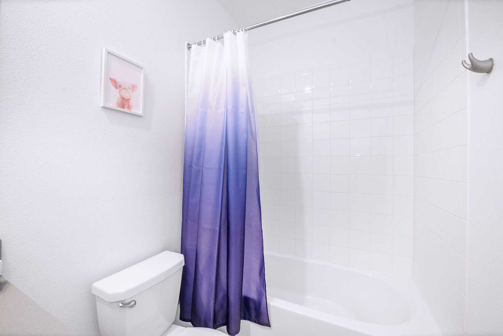 Bright bathroom with a white bathtub/shower combo, a distinctive purple ombre shower curtain, and a toilet. A small framed pig artwork adds a playful touch to the white walls.