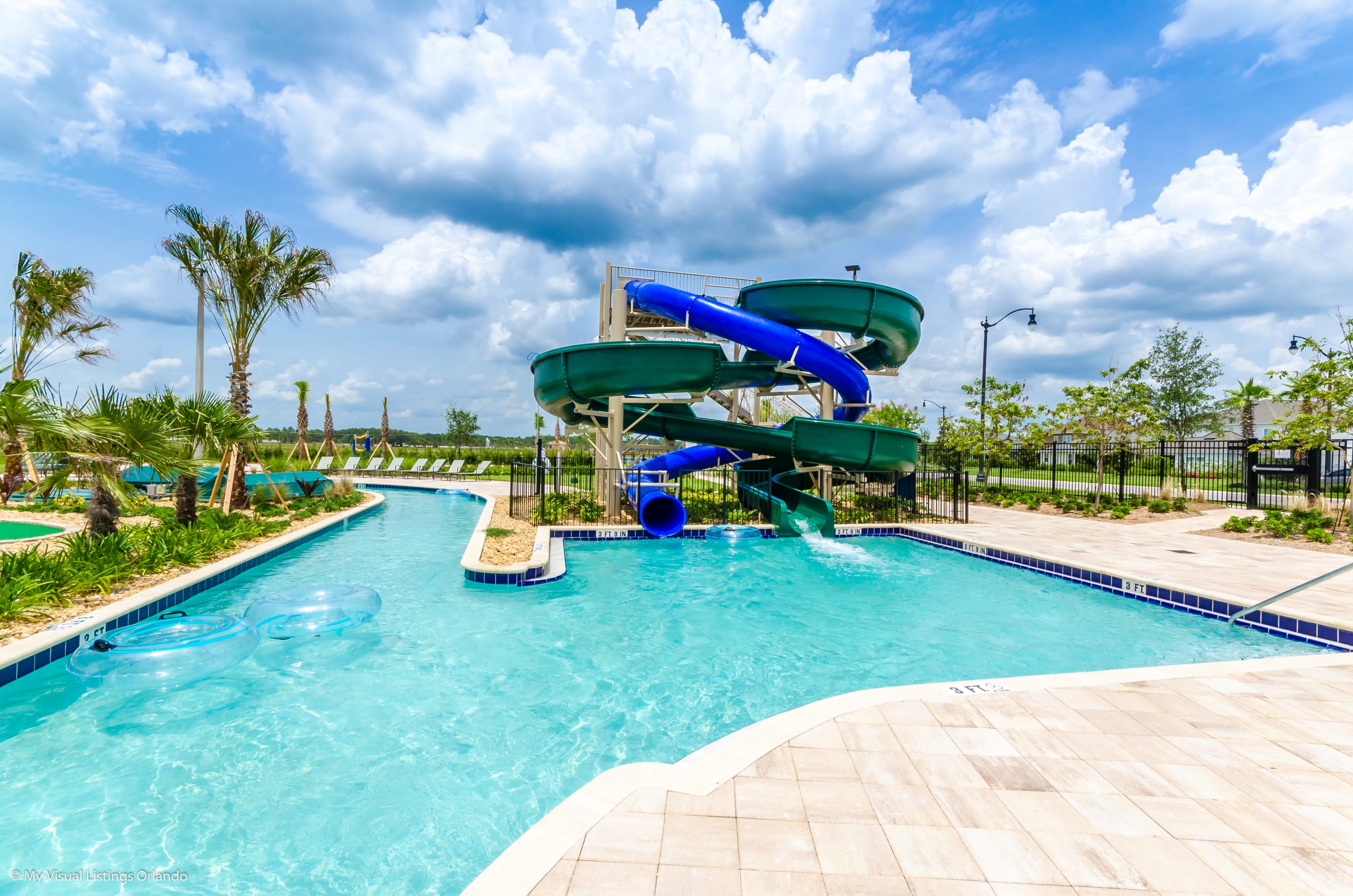A vibrant outdoor communal pool area features a lazy river with inner tubes and a multi-level water slide with green and blue enclosed flumes. Palm trees and lounge chairs line the pool's edge under a bright blue sky with fluffy white clouds.