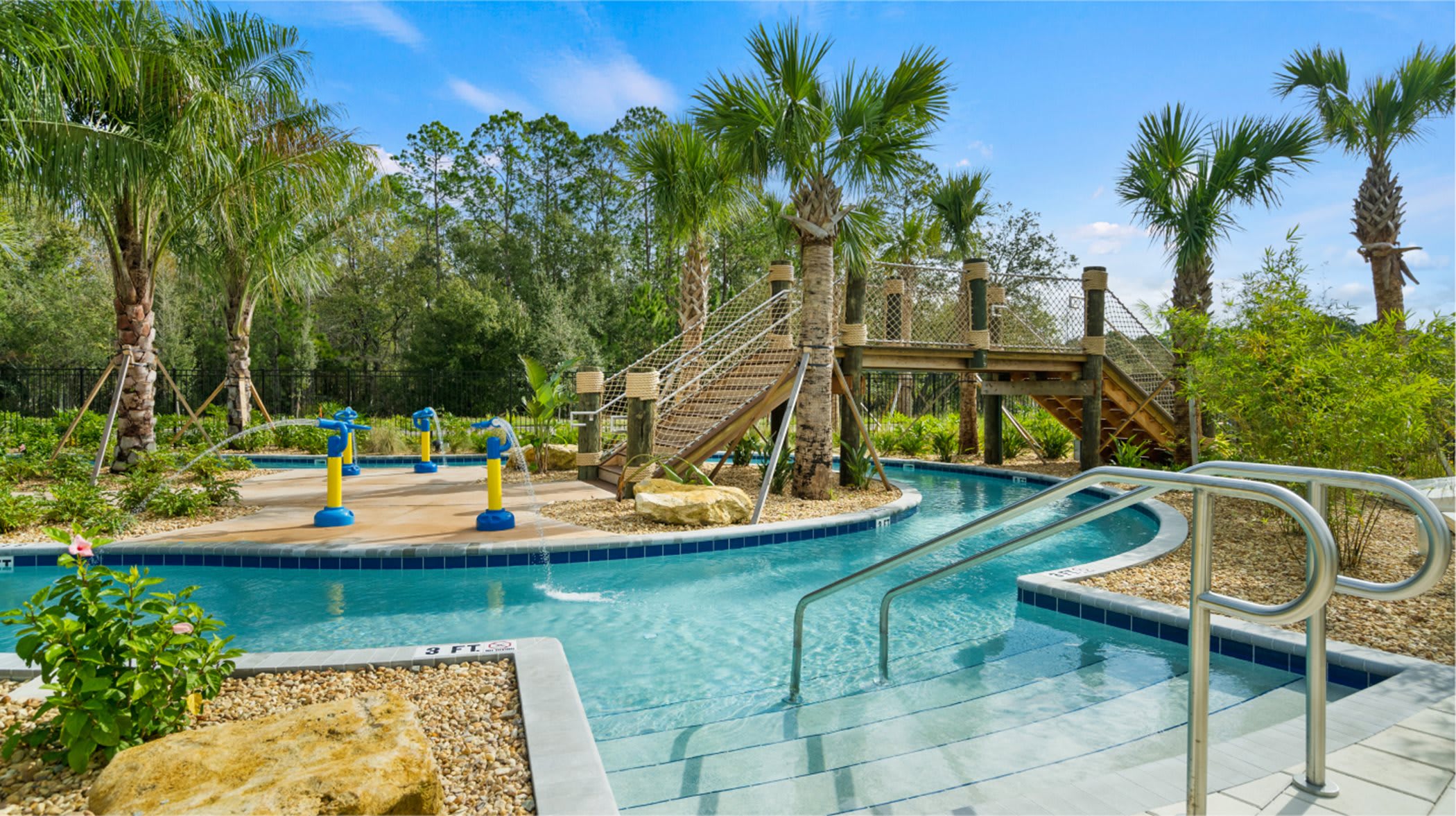 Inviting lazy river with steps leading into the water, featuring water sprayers and a rustic wooden bridge with rope accents, all set amidst vibrant green foliage and tall palm trees.