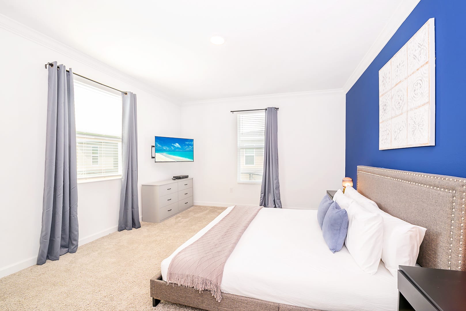Bright bedroom with a king bed, a blue accent wall, and two large windows with gray curtains, featuring a wall-mounted TV above a gray dresser, all on a light beige carpet.