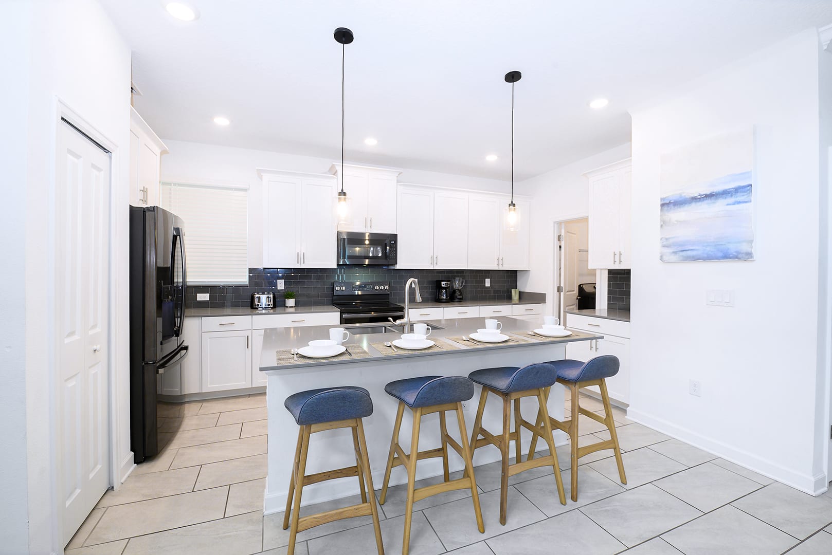 Modern kitchen with white shaker cabinets, dark gray subway tile backsplash, and light gray countertops. A large island with a double sink and blue bar stools serves as a breakfast bar, featuring stainless steel appliances.