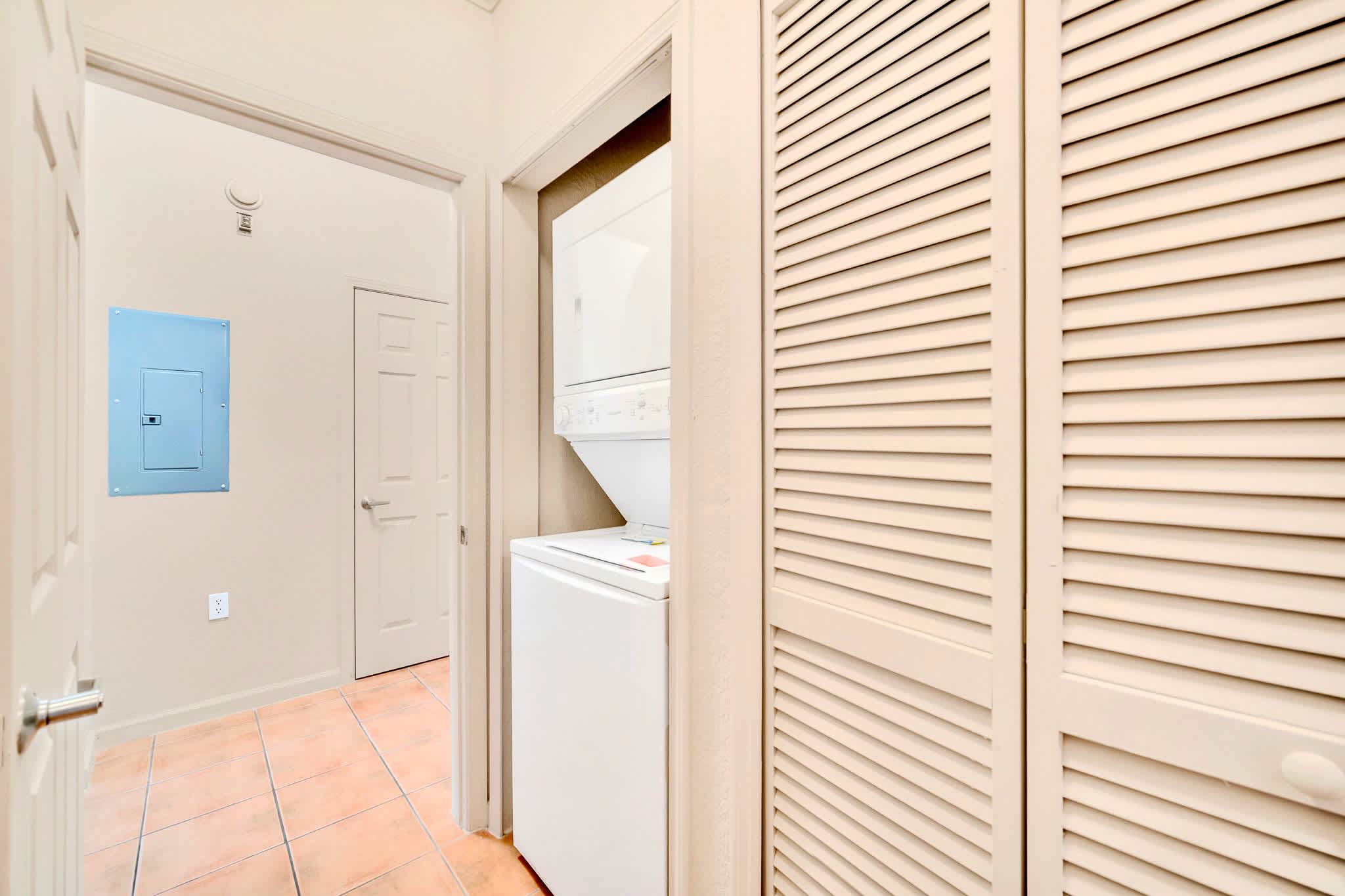A small laundry area with a stacked white washer and dryer unit, located in a hallway with light beige walls and terracotta-colored tile flooring.