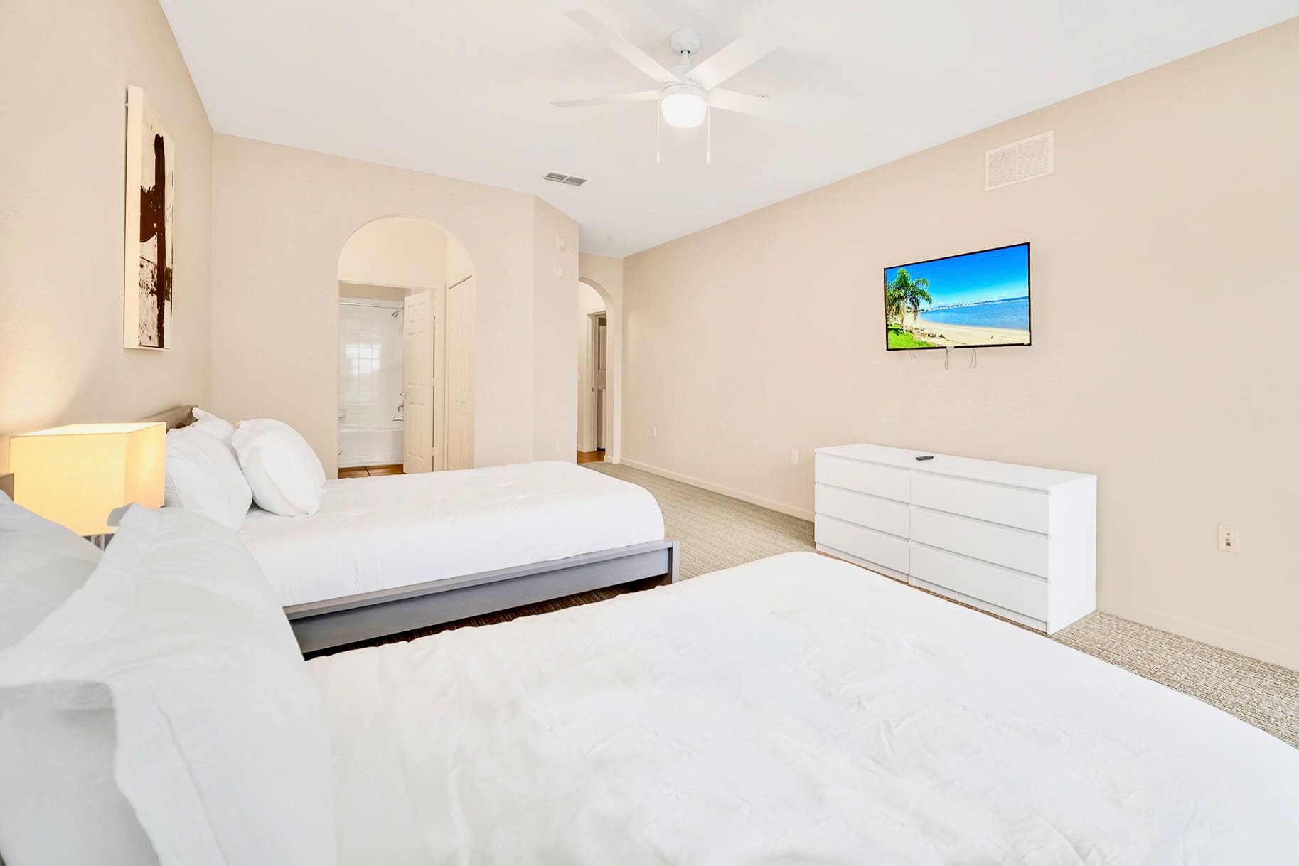 A wide shot of the second bedroom, featuring two twin beds with white bedding, a nightstand with a lamp between them, and a wall-mounted TV above a white dresser. An arched doorway leads to another room.