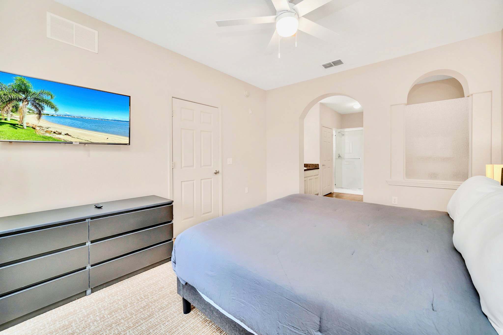A bright bedroom with light beige walls, a large gray bed with a dark gray comforter, a wall-mounted TV displaying a beach scene, and a dark gray dresser. An arched doorway leads to an ensuite bathroom.
