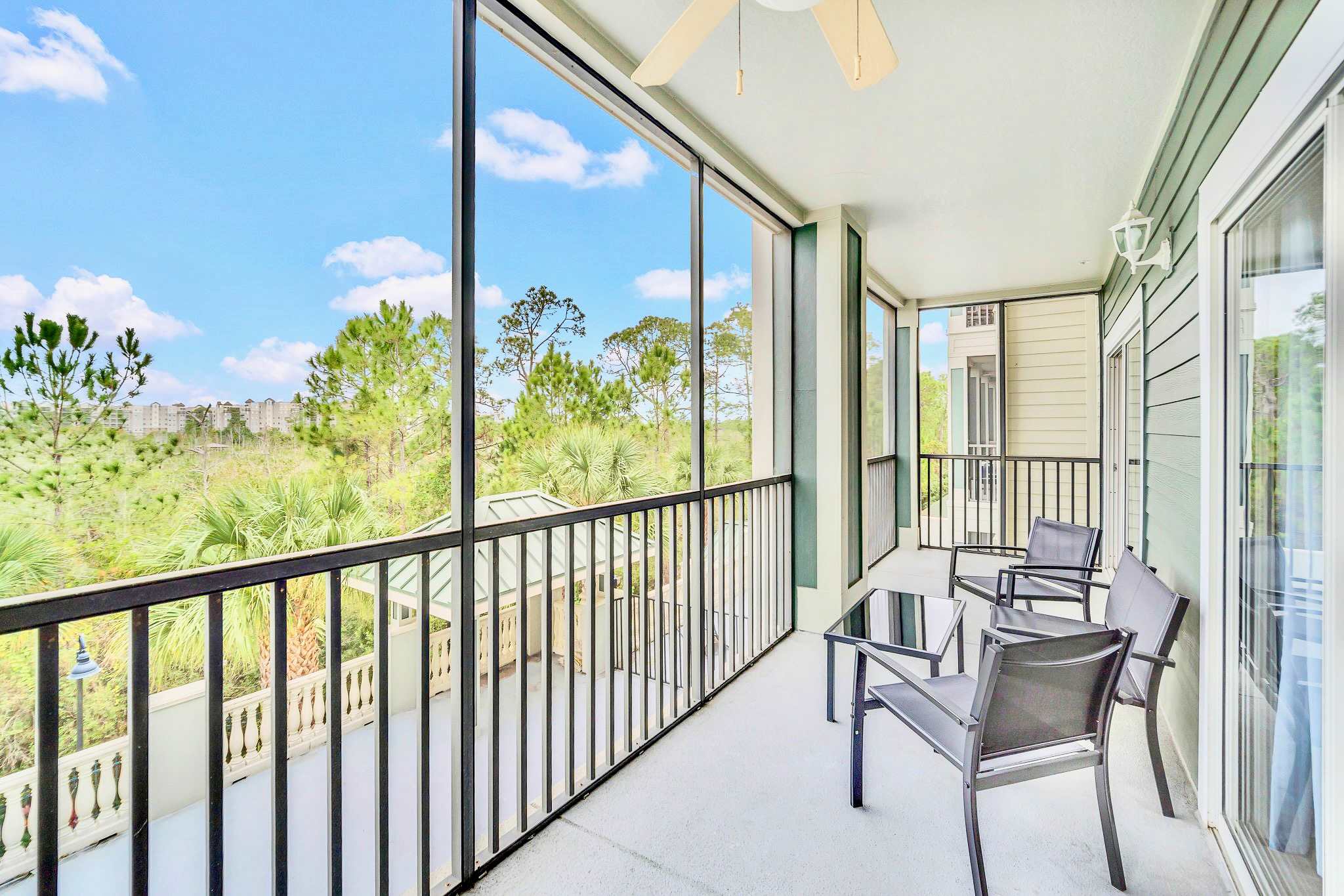A screened-in balcony with black metal railings, featuring outdoor seating including two chairs and a small table. It offers a view of lush green trees and distant buildings under a blue sky.