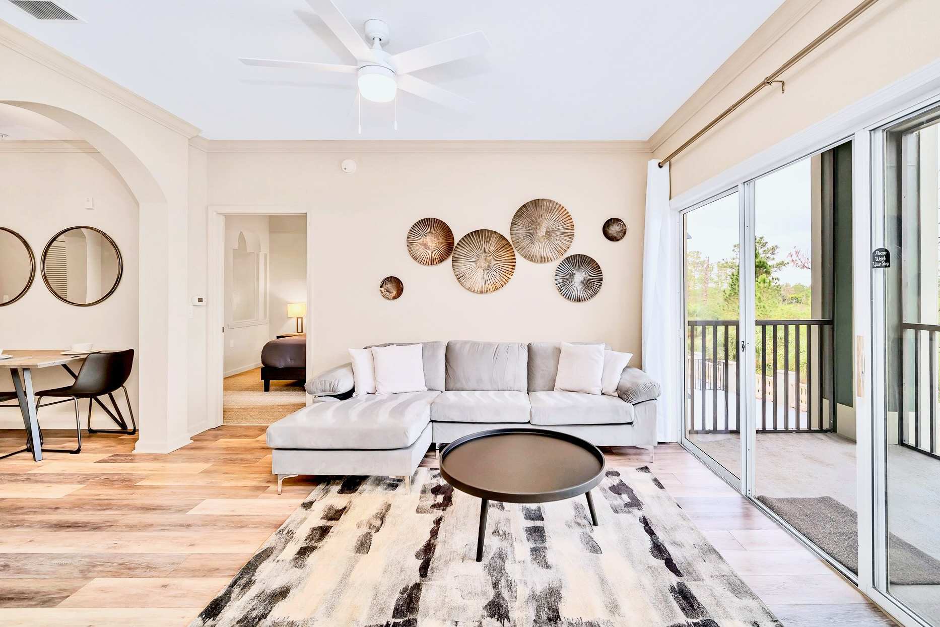 Inviting living room featuring a light gray sectional sofa, a dark round coffee table on a geometric rug, and a wall adorned with circular metallic art pieces, with balcony access visible.