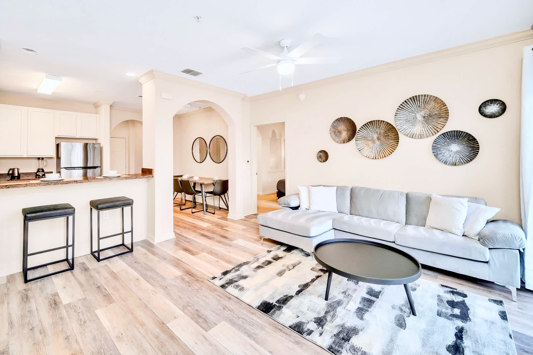 Open-concept living space showcasing a light gray sectional sofa on a patterned rug, a dark round coffee table, and a breakfast bar with two stools, leading into the dining area and kitchen.