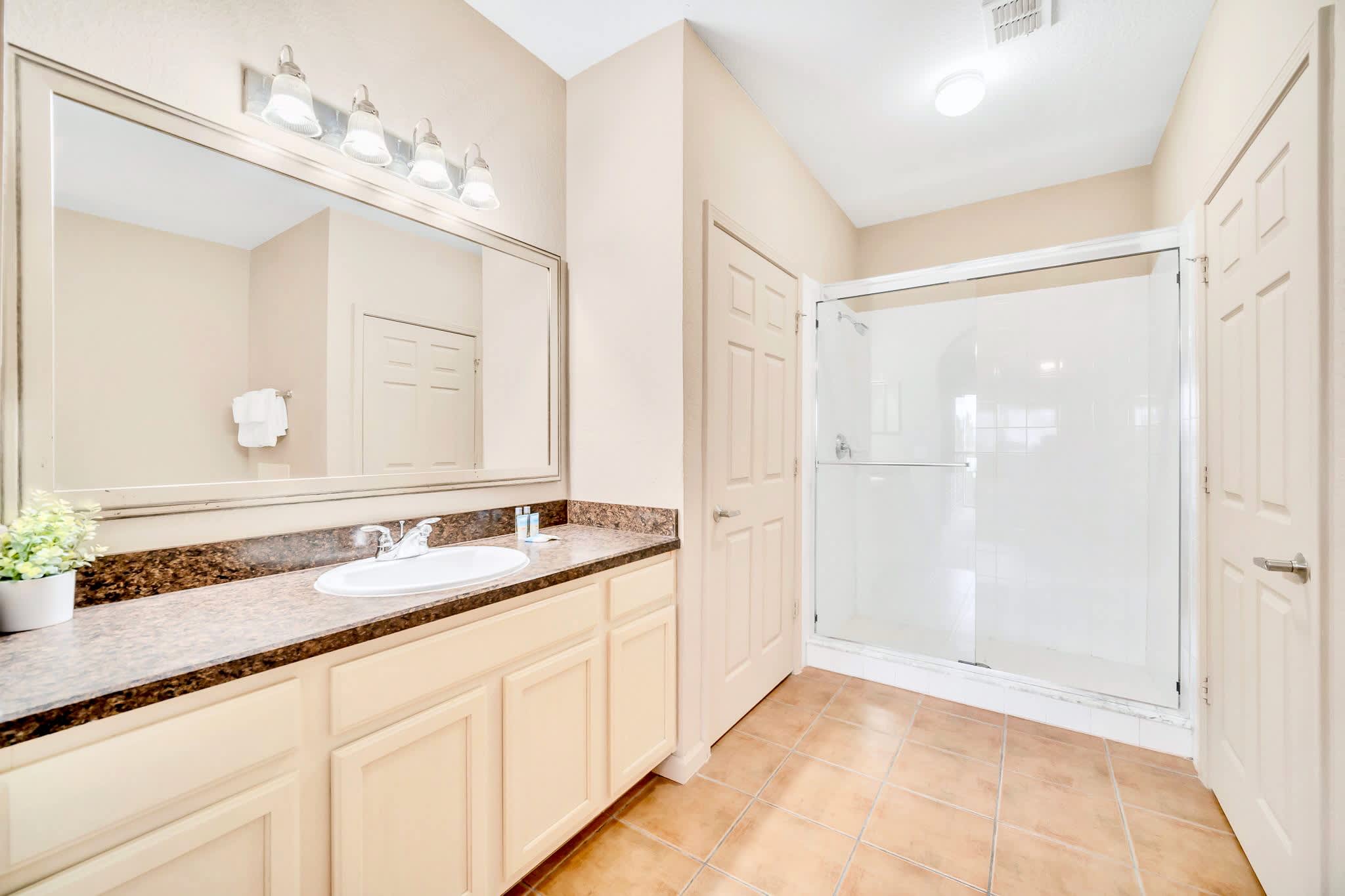 A spacious bathroom with light beige walls, a large vanity with a white sink, dark speckled countertop, and light cabinets. It features a large mirror with vanity lighting and a glass-enclosed walk-in shower.
