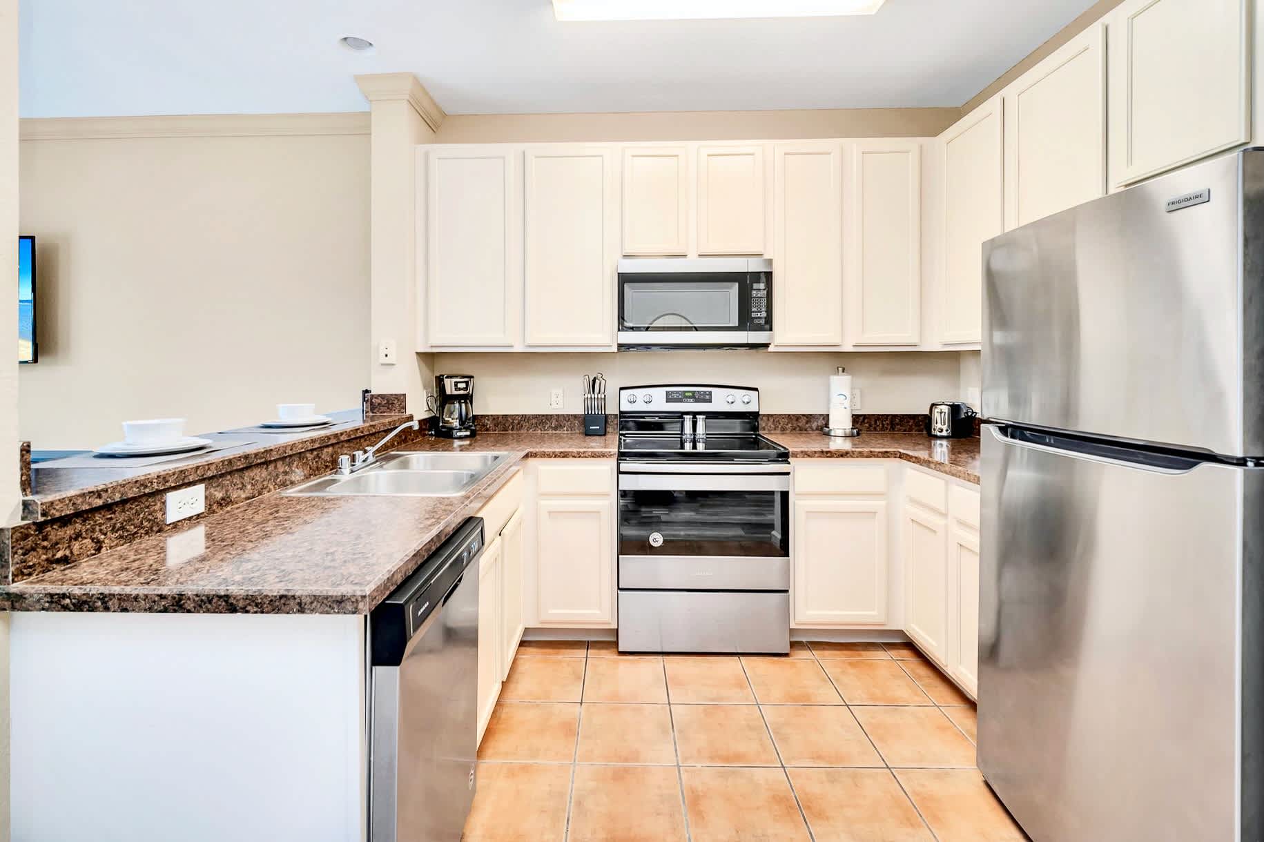 Fully equipped kitchen with light cream upper and lower cabinets, speckled granite countertops, and stainless steel appliances including a refrigerator, stove, and microwave, set against terracotta-colored tile flooring.