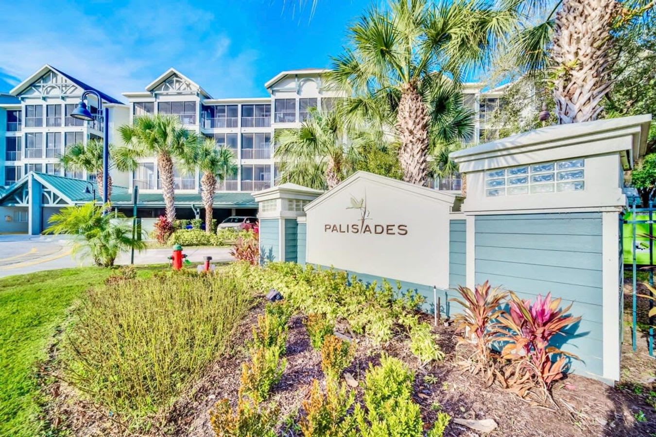 Front exterior view of a multi-story building with light blue and white siding, surrounded by lush green landscaping and palm trees, featuring a "PALISADES" sign at the entrance.
