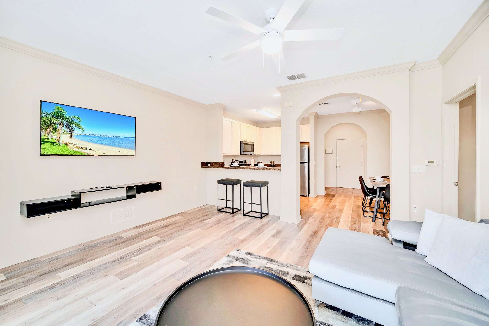 Modern living room with a wall-mounted television above a floating black media console, facing a breakfast bar with two stools, and an open view towards the kitchen and dining areas.