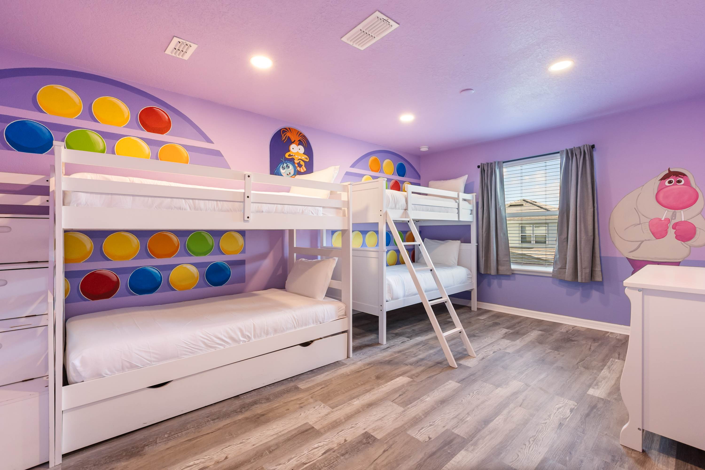 A colorful kids' bedroom with a vibrant purple theme and "Inside Out" character murals on the walls. Two white bunk beds with trundles are positioned against the walls, complemented by wood-look flooring.