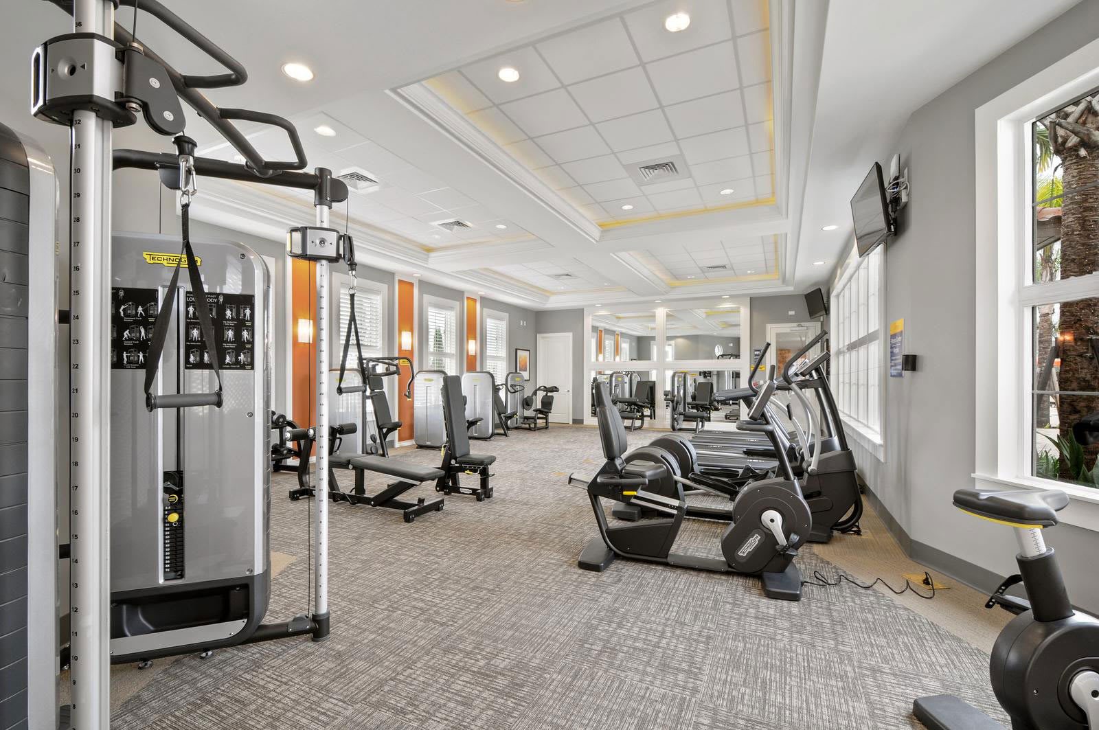 A well-equipped indoor gym features a variety of modern exercise machines including treadmills, stationary bikes, and weight machines, set on a patterned carpet with large windows and bright overhead lighting.