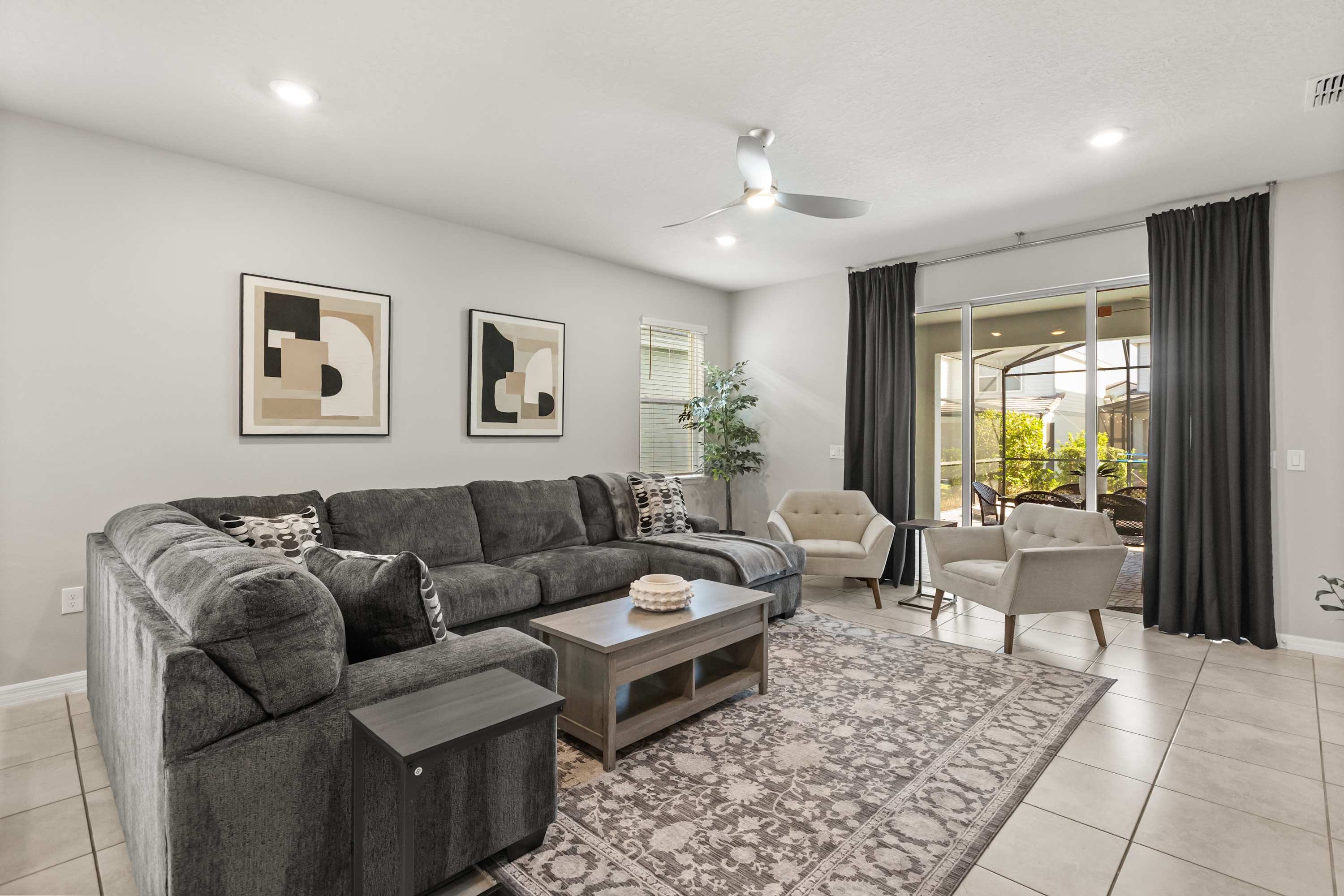 Spacious living room featuring a large gray sectional sofa, a light wood coffee table, and a patterned area rug on light tile floors. Sliding glass doors open to an outdoor patio, and two abstract art pieces adorn the light gray wall.