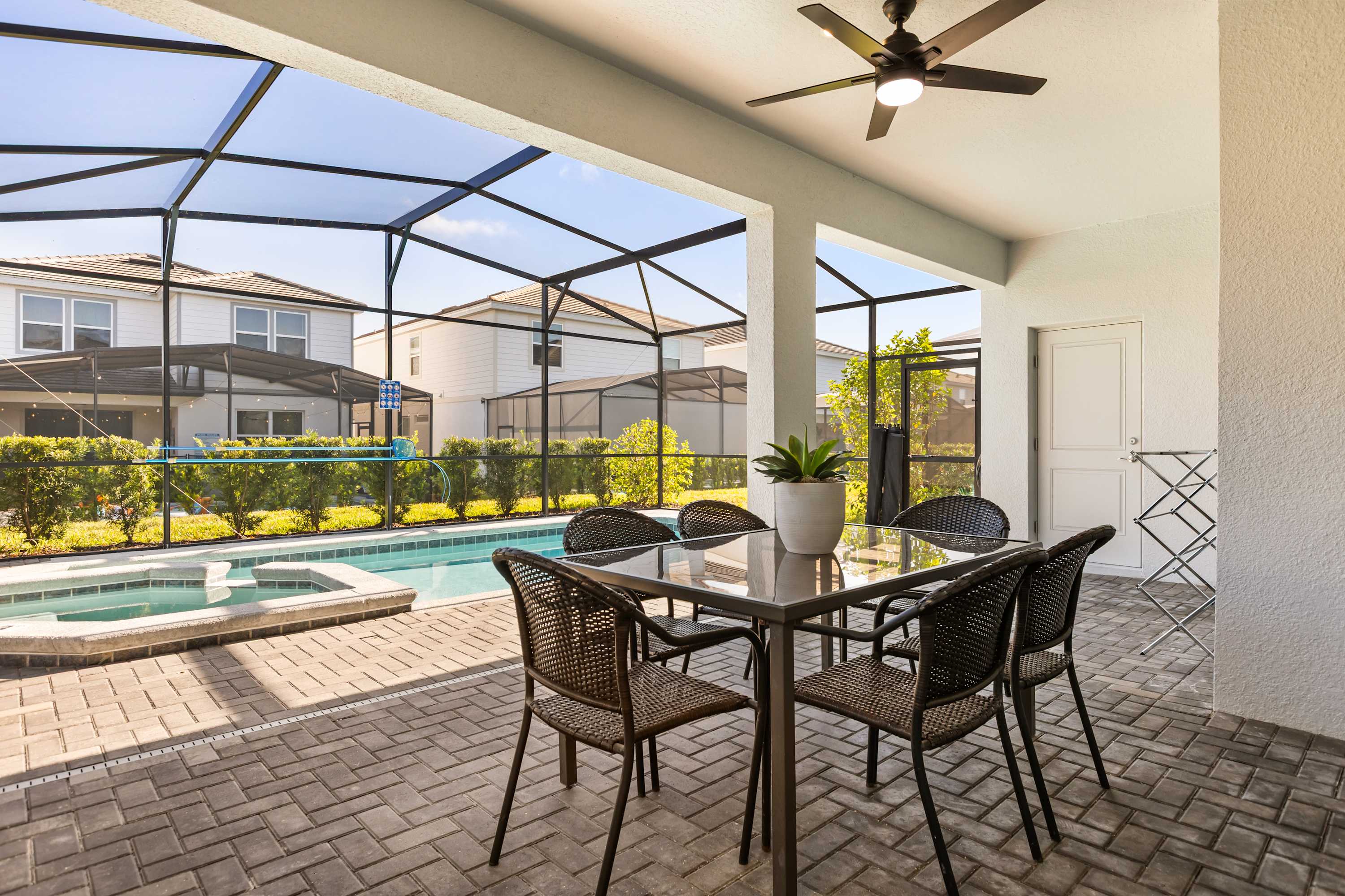 A covered outdoor patio features a dark square dining table with six wicker chairs and a ceiling fan, overlooking a screened-in pool and hot tub area. The space is paved with light-colored bricks.
