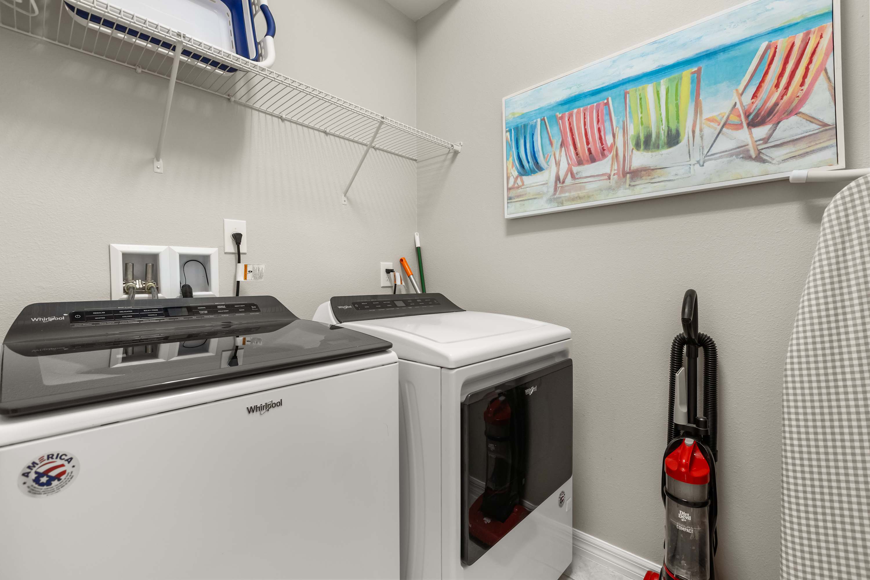 A functional laundry room equipped with a white top-load washer and front-load dryer, a wire shelf for storage, and light gray walls. A colorful beach-themed artwork hangs above the appliances, and a vacuum cleaner stands in the corner.