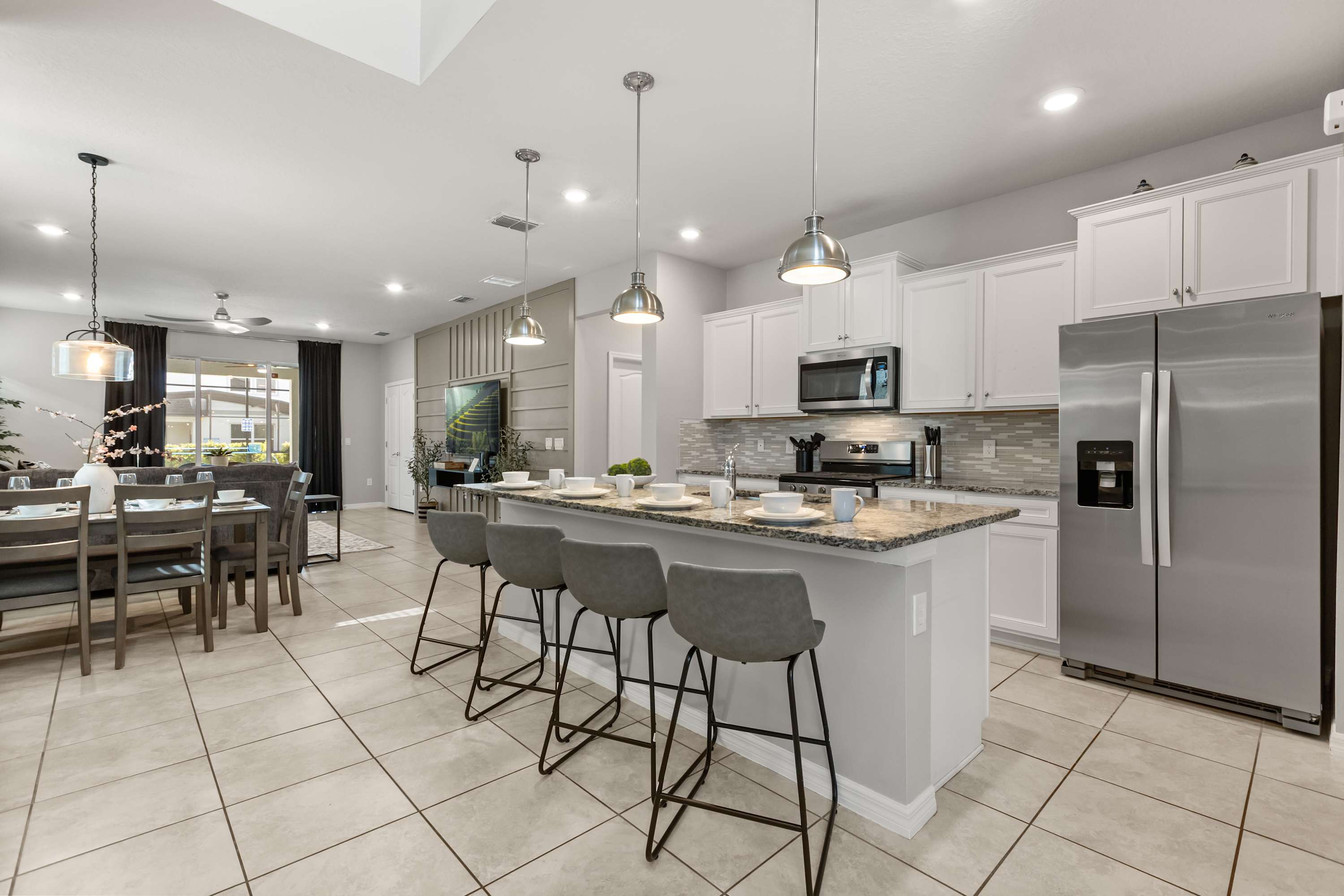Modern kitchen with white shaker cabinets, granite countertops, and stainless steel appliances, including a large refrigerator and microwave. A kitchen island with pendant lights and four gray bar stools separates the kitchen from the dining area.