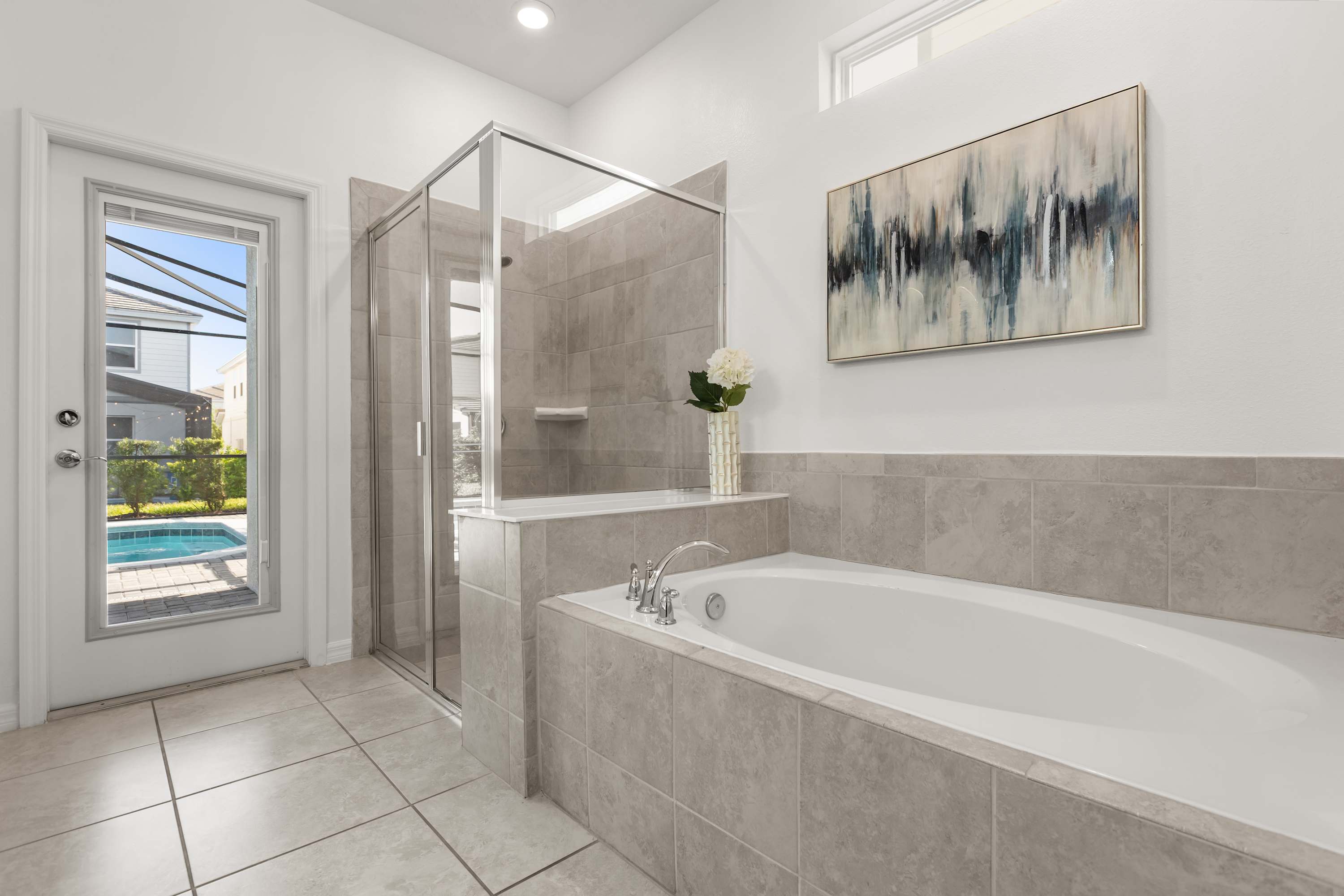 A spacious bathroom featuring a built-in soaking tub with tile surround and a separate glass-enclosed walk-in shower. A large abstract painting hangs above the tub, and a glass door provides access to the outdoor pool area.