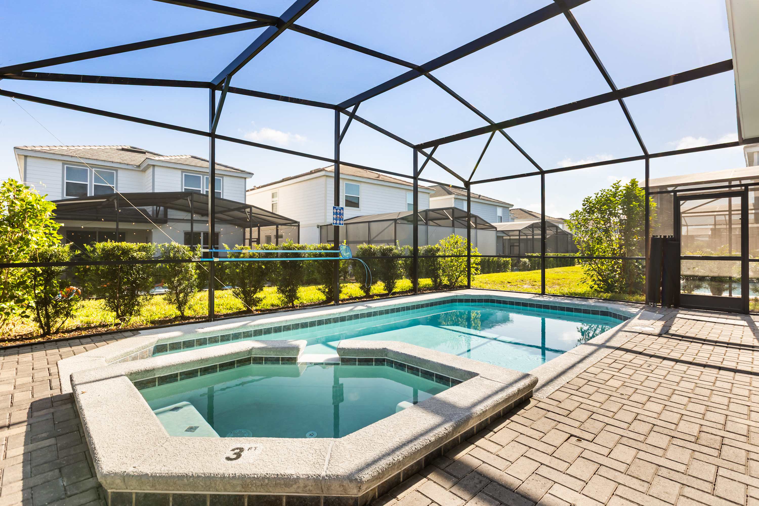 A bright outdoor screened-in pool area features a rectangular swimming pool and an adjacent hot tub, surrounded by light-colored brick pavers. The enclosure provides a clear view of neighboring houses and landscaping.