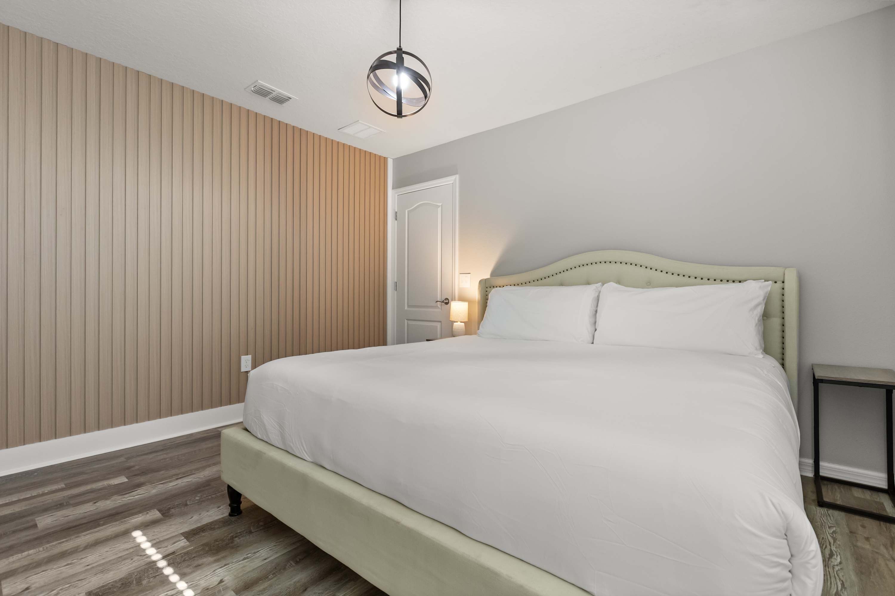 A serene bedroom with a large bed featuring a light upholstered, tufted headboard, white bedding, and light wood-look flooring. A modern spherical chandelier hangs from the ceiling, and a light wood slatted accent wall adds texture behind the bed.