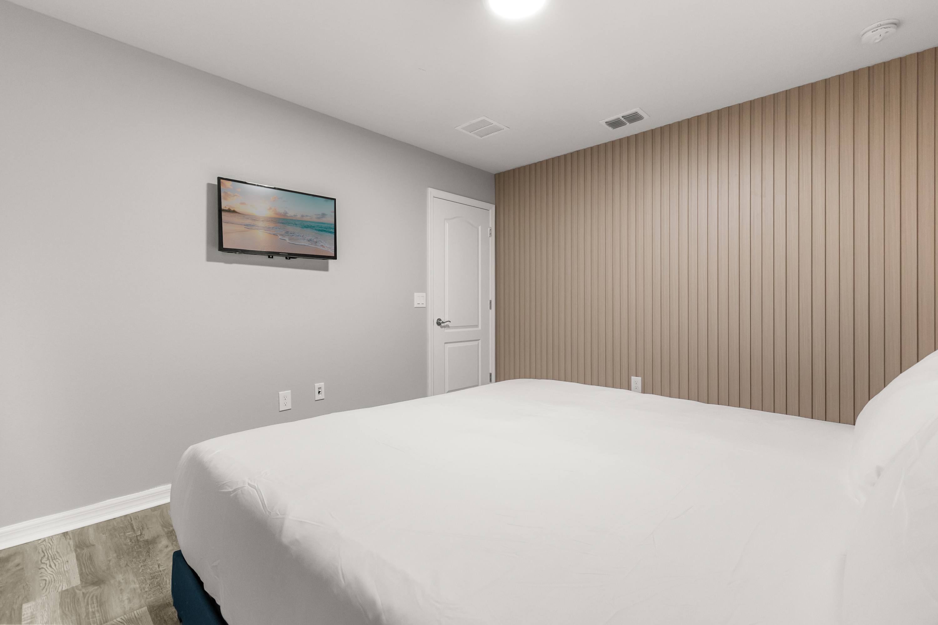 A bright bedroom featuring a large bed with white bedding, light gray walls, and a textured light wood accent wall behind the bed. A wall-mounted TV displays a beach scene, and light wood-look flooring completes the modern aesthetic.