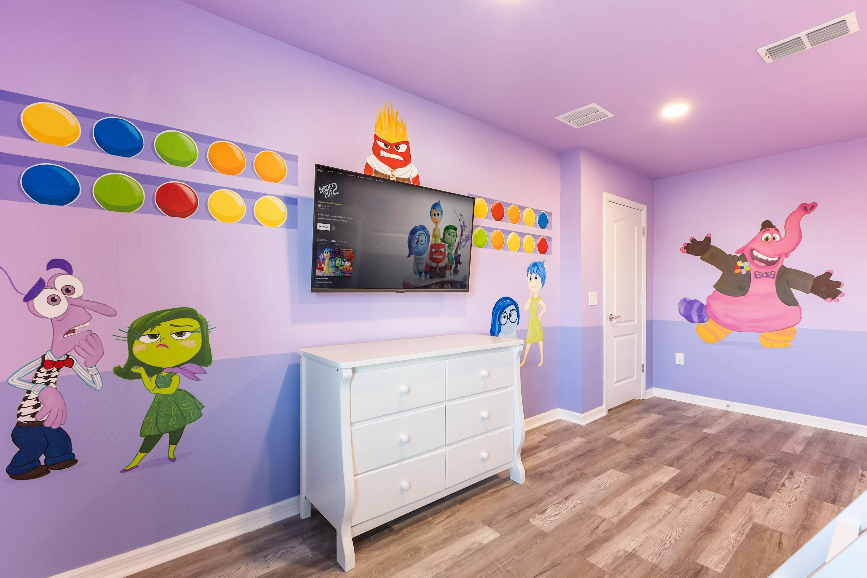 A detailed view of the "Inside Out" themed bedroom, showcasing various character murals on the purple walls and a wall-mounted TV above a white dresser. Colorful circular accents adorn the walls, adding to the playful atmosphere.