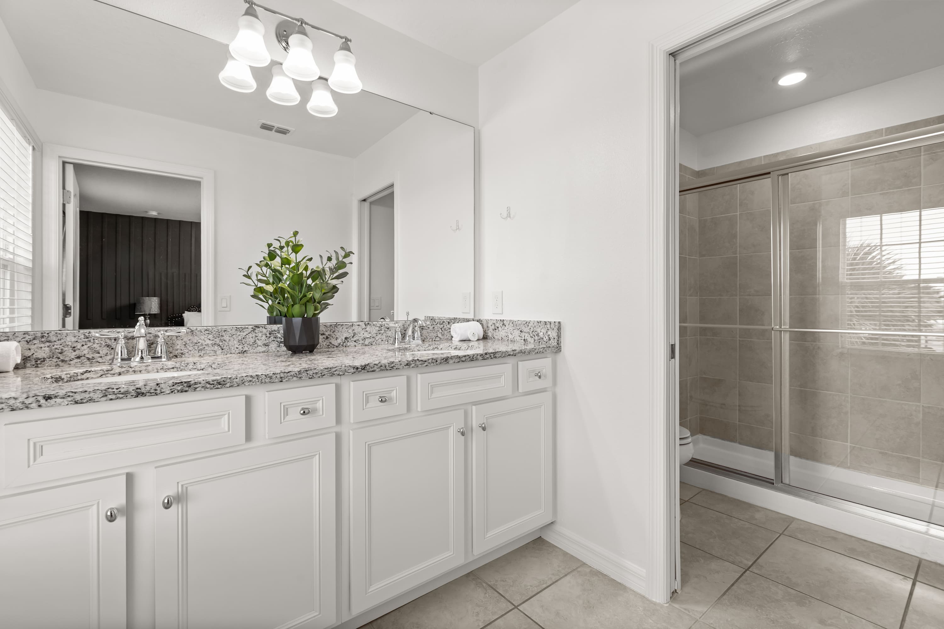 A bright bathroom featuring a double vanity with white cabinets, granite countertops, and two sinks. A large mirror spans the width of the vanity, and a glass-enclosed shower with tiled walls is visible to the right.