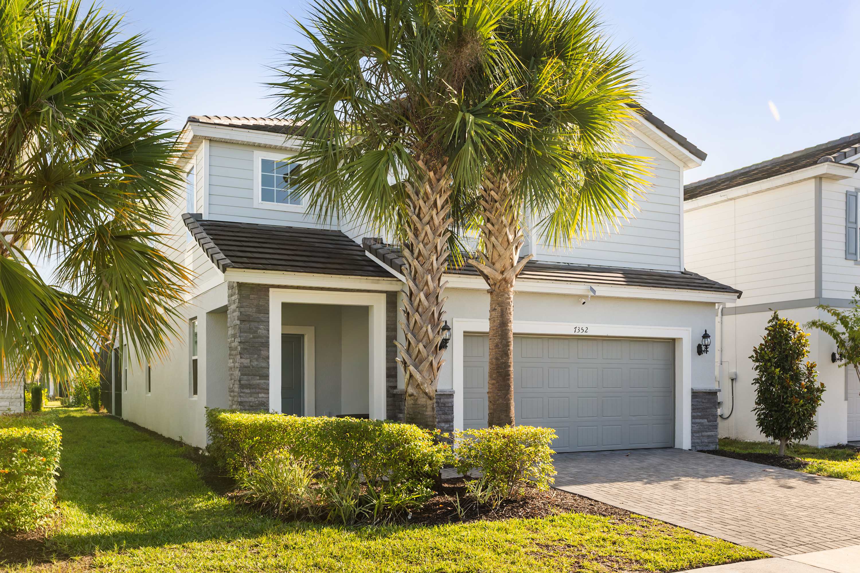 The front exterior of a two-story light blue house with gray stone accents and a gray garage door, set against a clear blue sky. Two tall palm trees and manicured bushes frame the entrance, with a paved driveway leading to the garage.