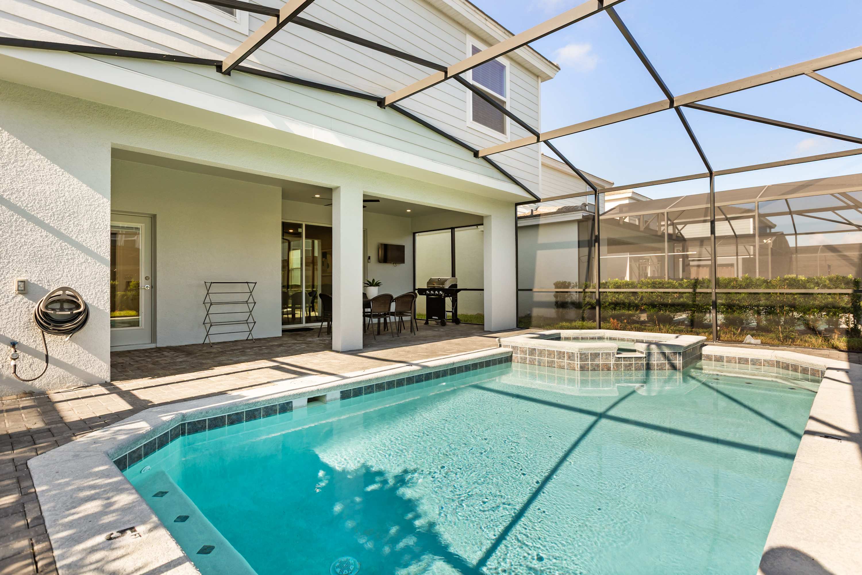 An expansive screened-in outdoor area features a rectangular swimming pool and hot tub, with a covered patio section offering shade. The patio includes an outdoor dining set and a BBQ grill, all surrounded by brick pavers.
