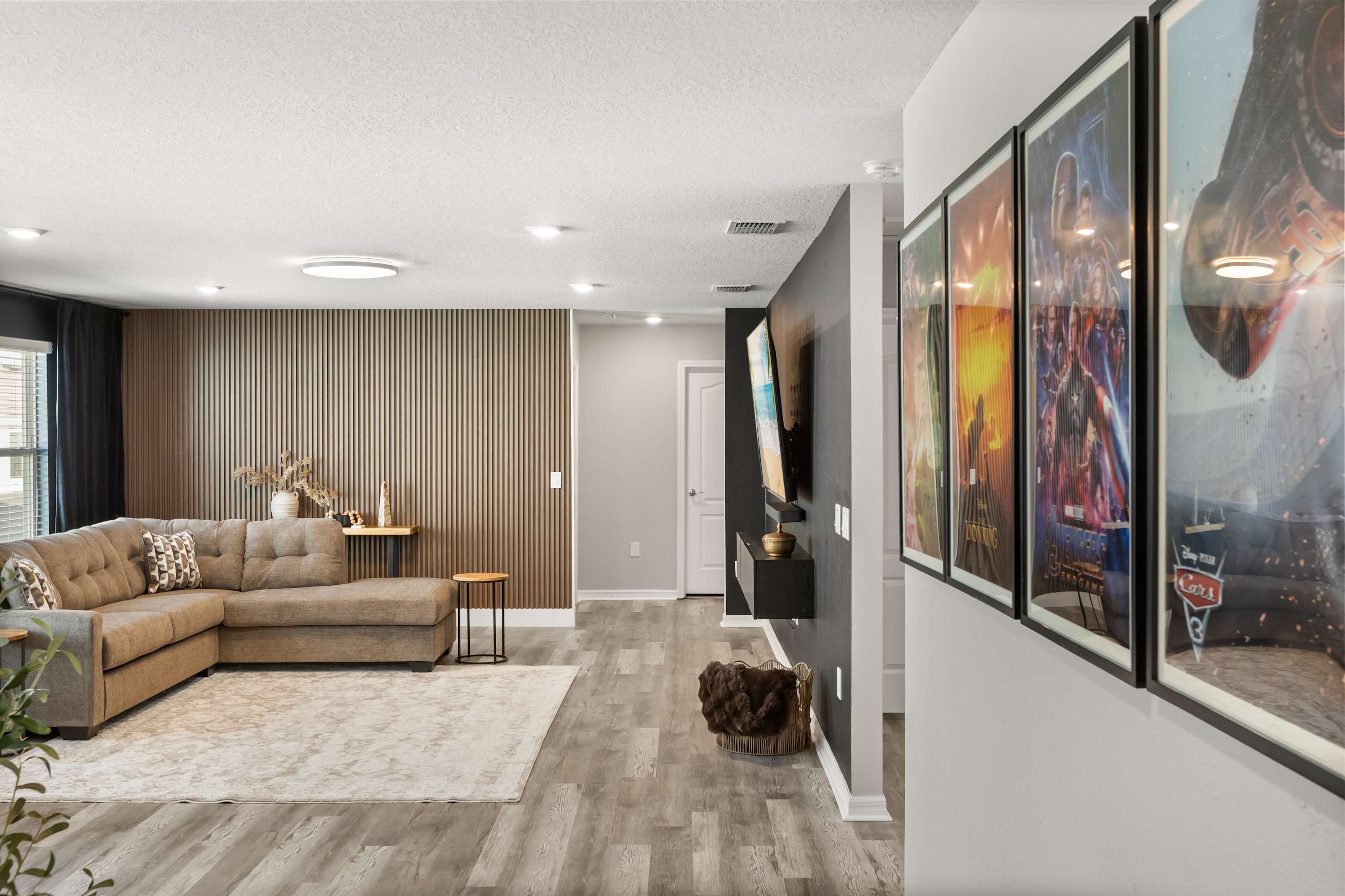 A spacious living area features a large L-shaped sectional sofa, a light-colored area rug, and a modern slatted accent wall. Framed movie posters adorn a nearby wall, leading to another section of the house.
